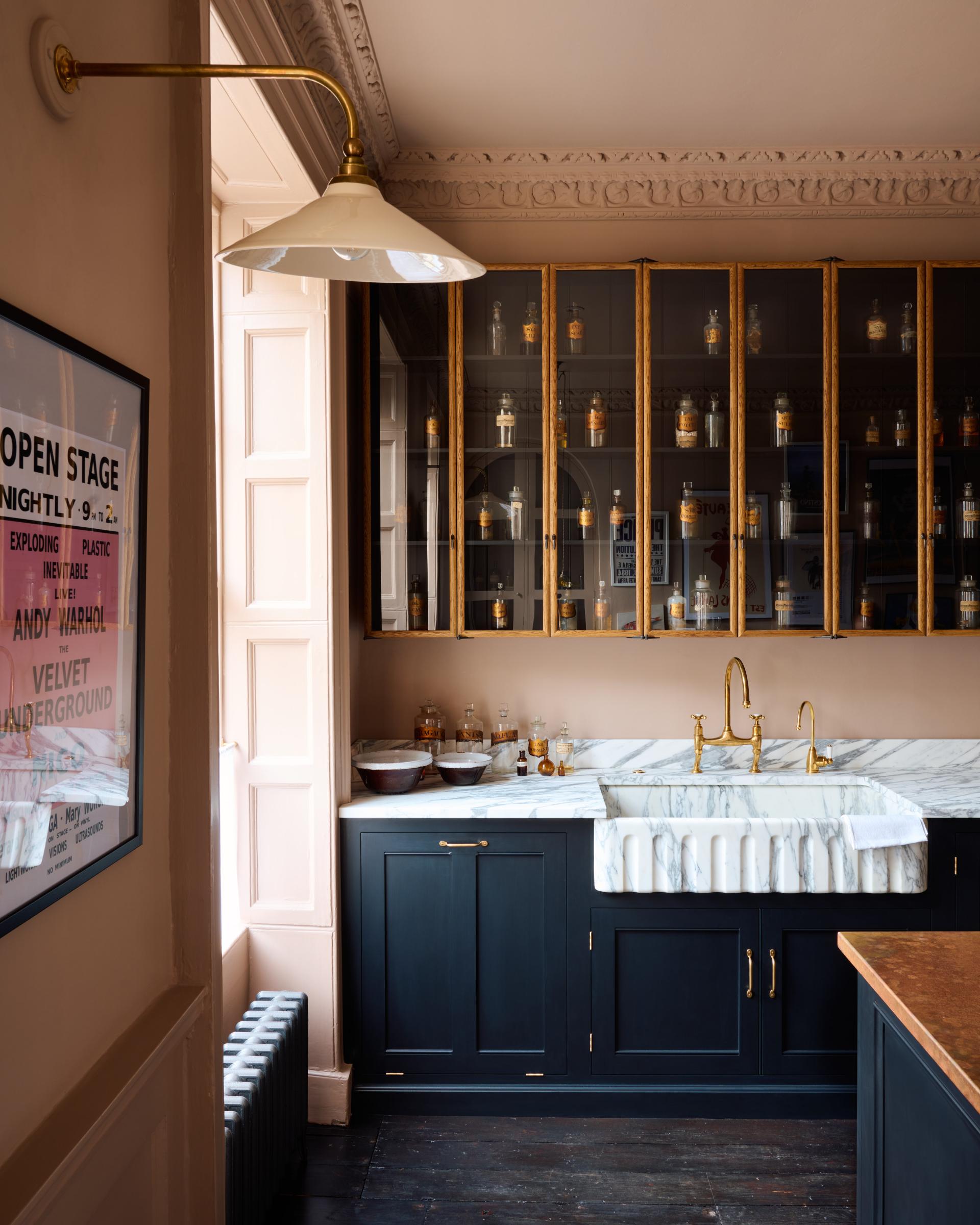 A straight-on view of a black painted Georgian-style kitchen in an atmospheric room with dark floorboards, pink walls and tall mid-century-style glazed oak wall cupboards.