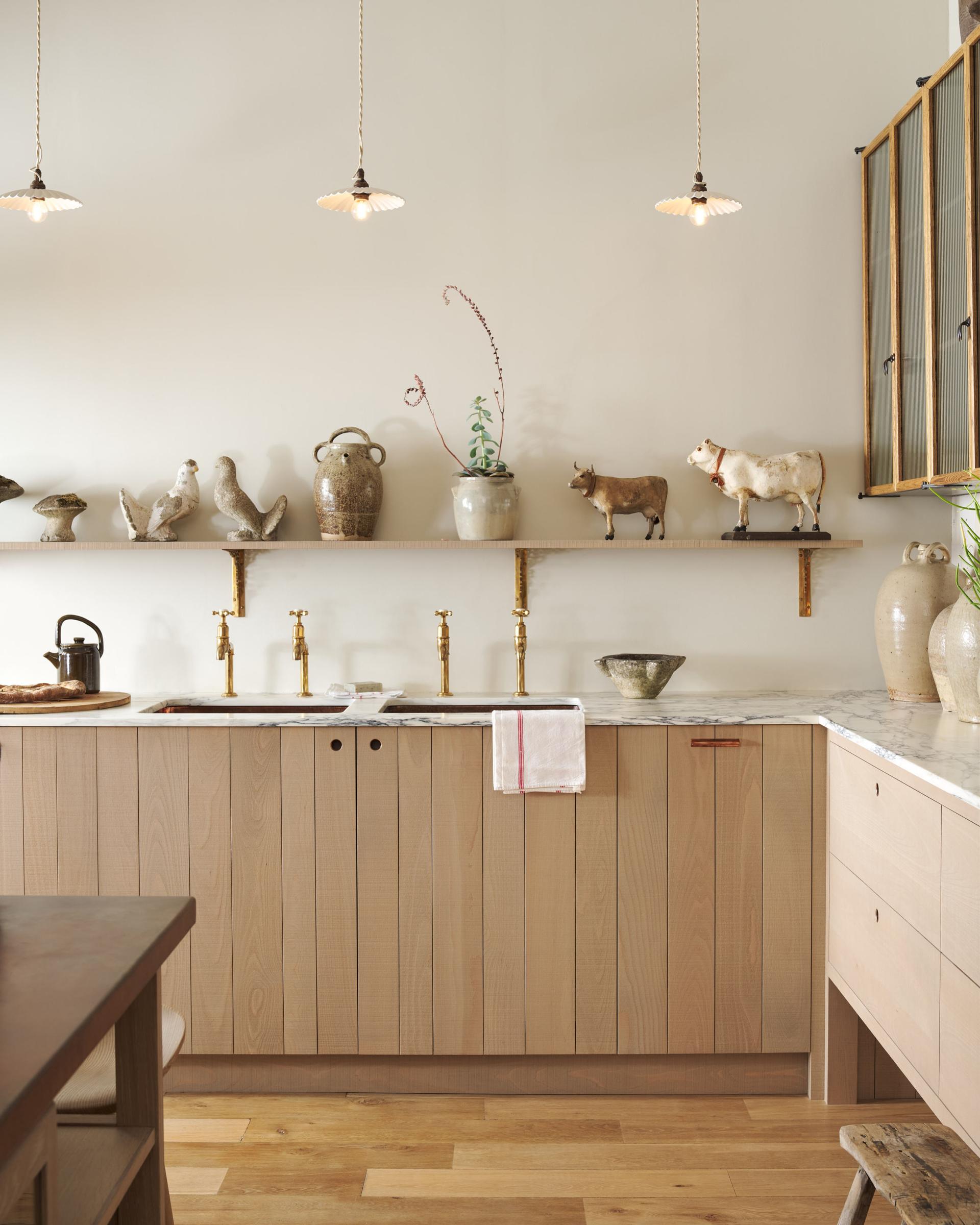 An all-natural and functional Sebastian Cox kitchen features an L-shaped sink run with a marble worktop, glazed wall cupboards, and an iconic Haberdasher’s prep table with a copper worktop. Lovingly adorned with antique artisan pottery, ceramic pendant lights, beautiful houseplants, and beloved figurines, it creates a wonderfully simple kitchen with a love of natural sustainability.
