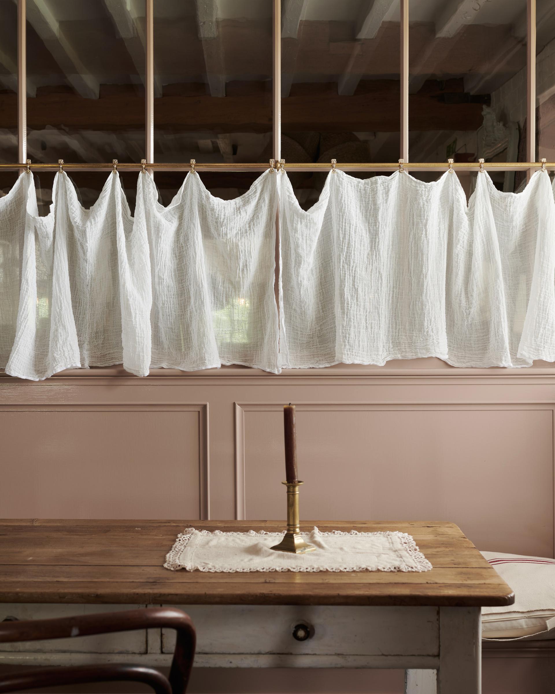 A straight-on view of a traditional pink painted glazed partition wall with an aged brass curtain rail attached to the central horizontal frame. A delicate linen curtain is clipped in several places along the length of the rail.
