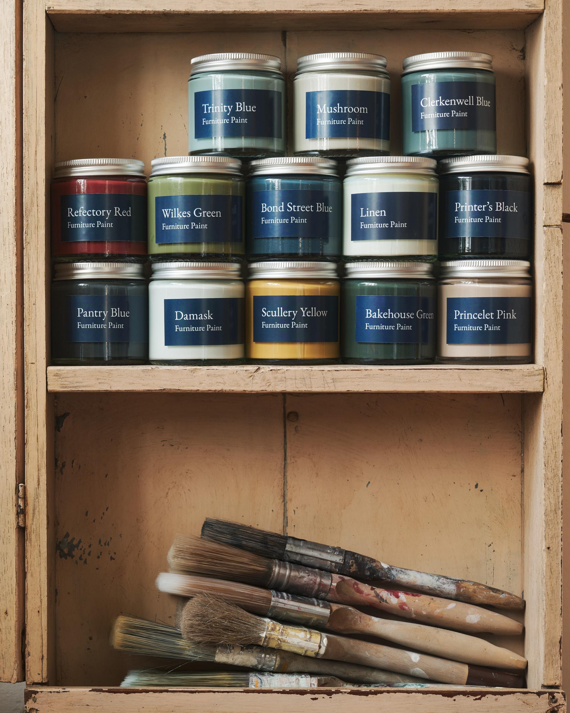A collection of glass jars with silver lids and blue labels in a vintage cupboard, each jar contains a different colour paint. A stack of worn paintbrushes can be seen on the shelf below.