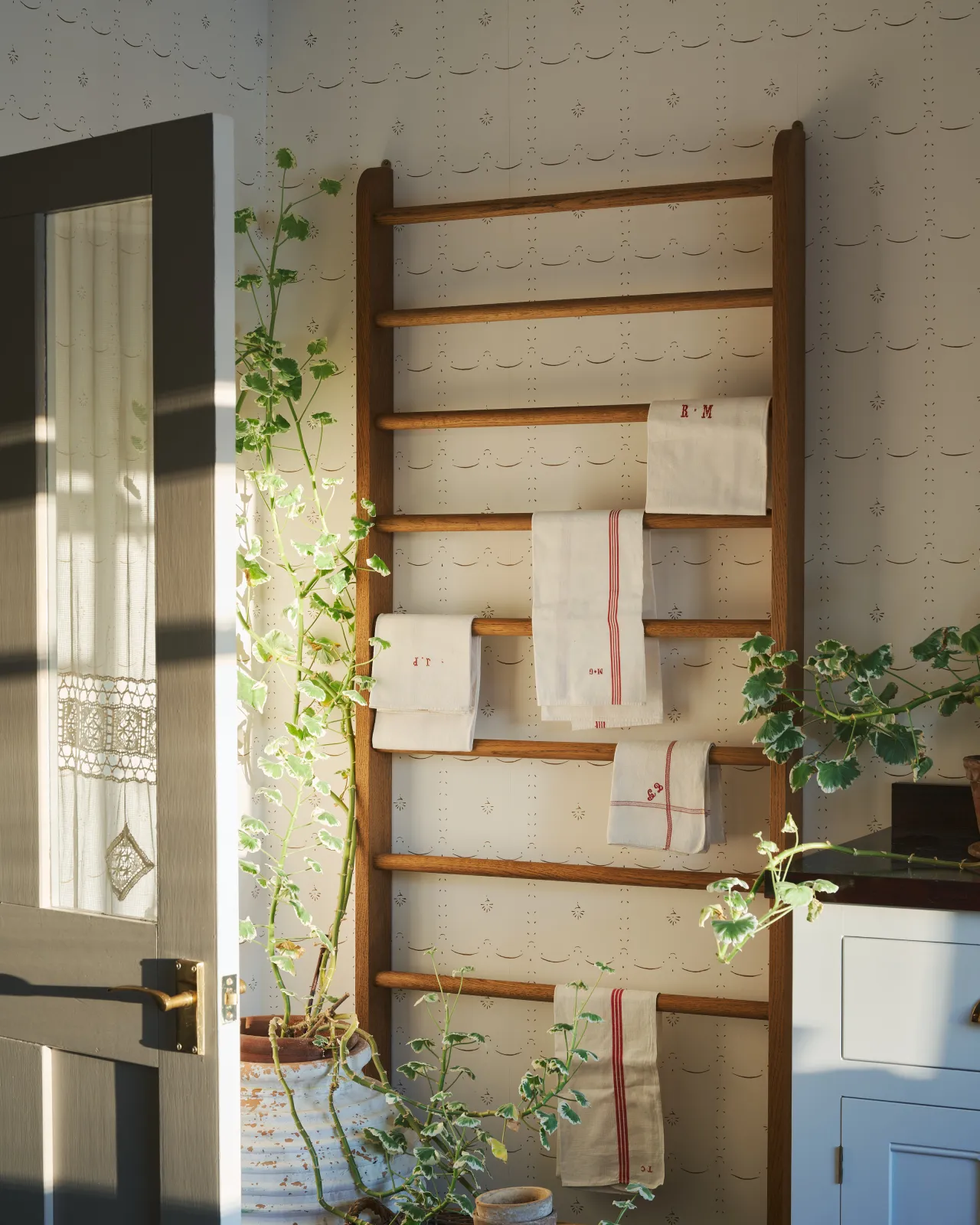 Strong light pours across a delicate patterned wallpaper behind a mid-century-style oak hanging rack, which is covered in vintage tea towels.