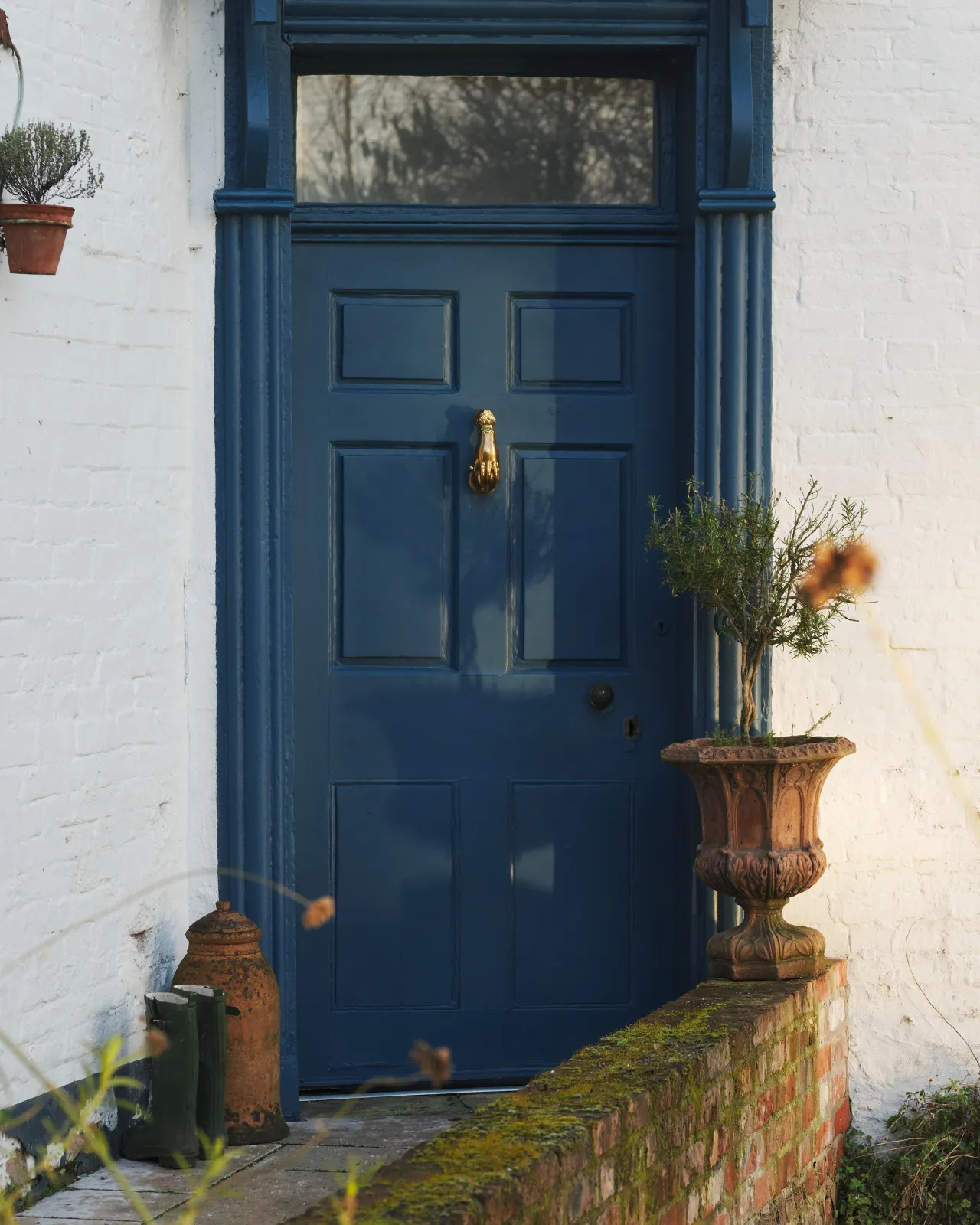 A straight-on view of a cottage with white brick walls and a blue painted front door and a decorative brass door knocker, shaped like a hand holding a pomegranate.