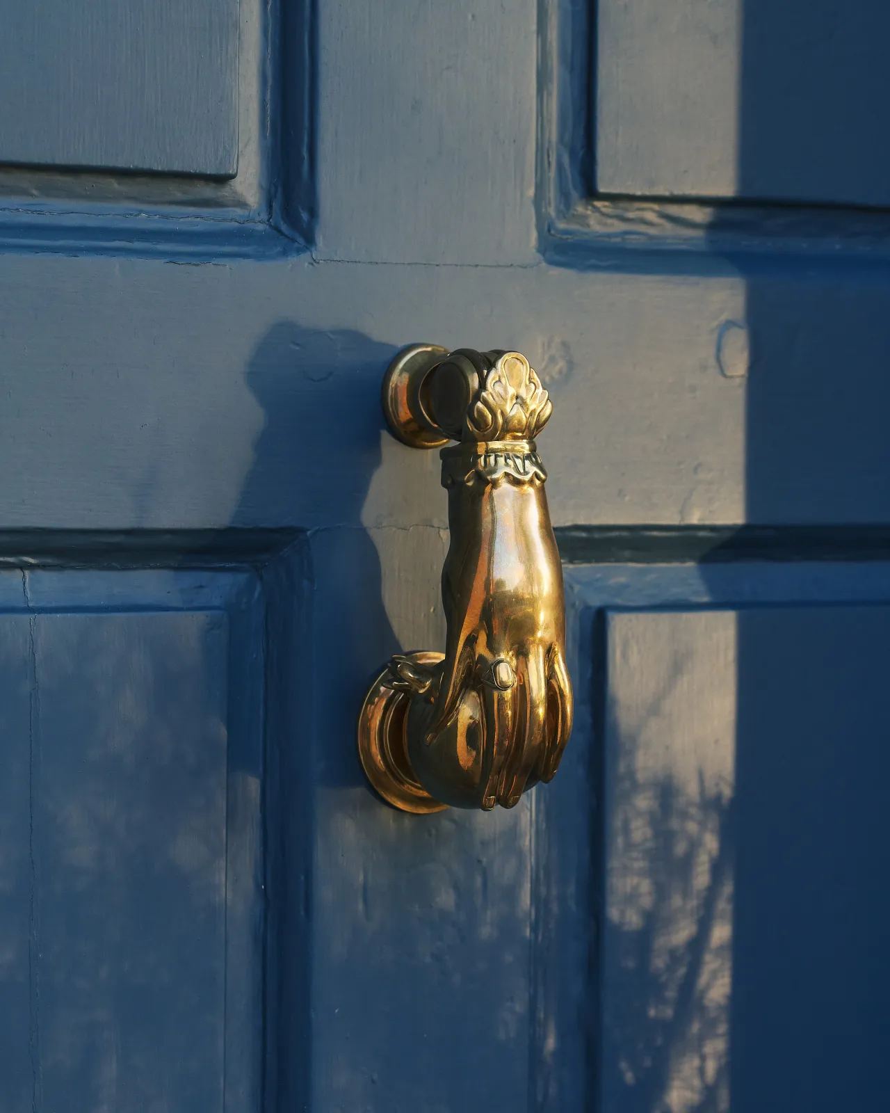 A close-up oblique view of a decorative brass door knocker, shaped like a hand holding a pomegranate and fitted to a blue painted door.