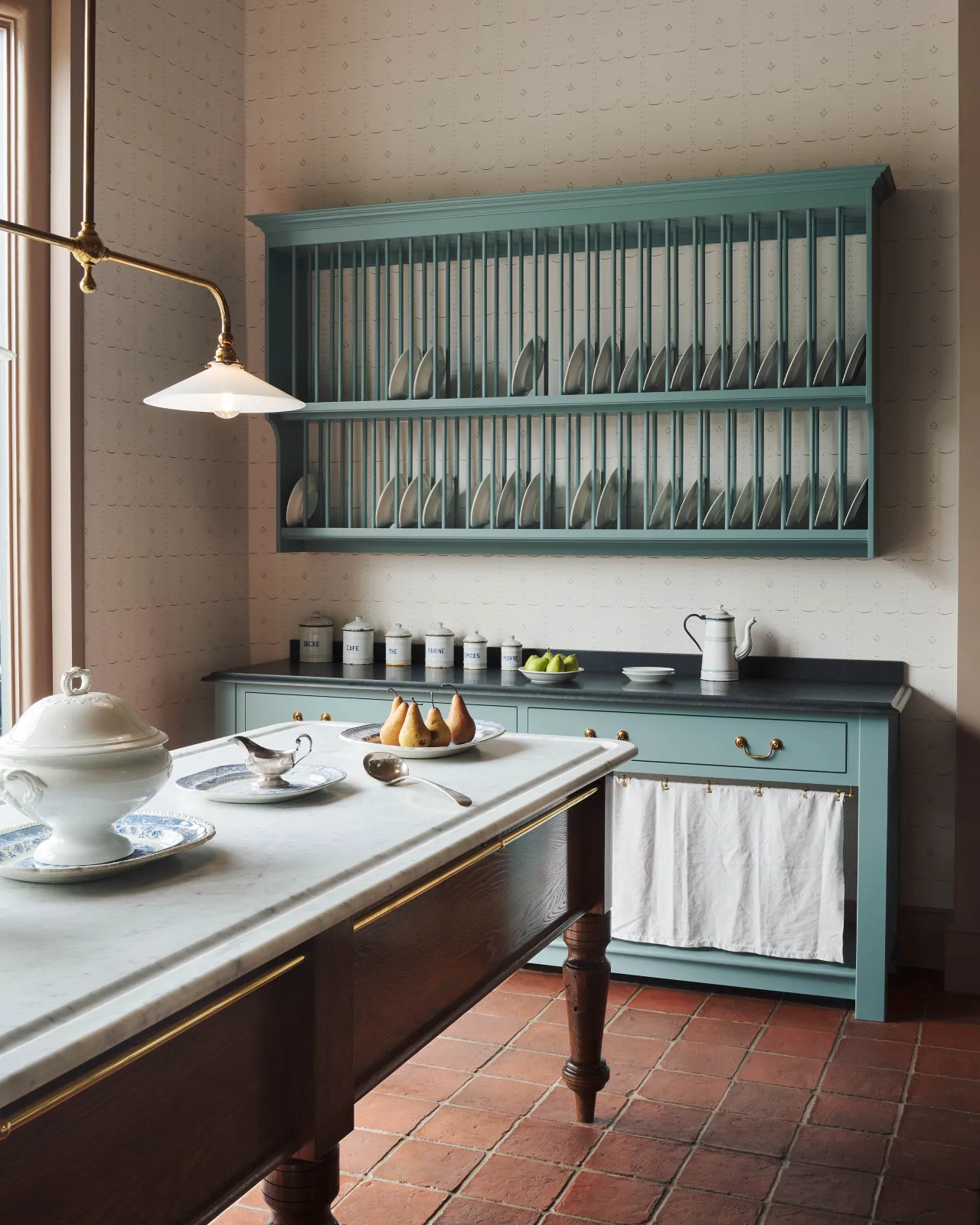 A Victorian-style English kitchen with classic cabinetry, a wall plate rack, large prep table and a rustic terracotta tiled floor.