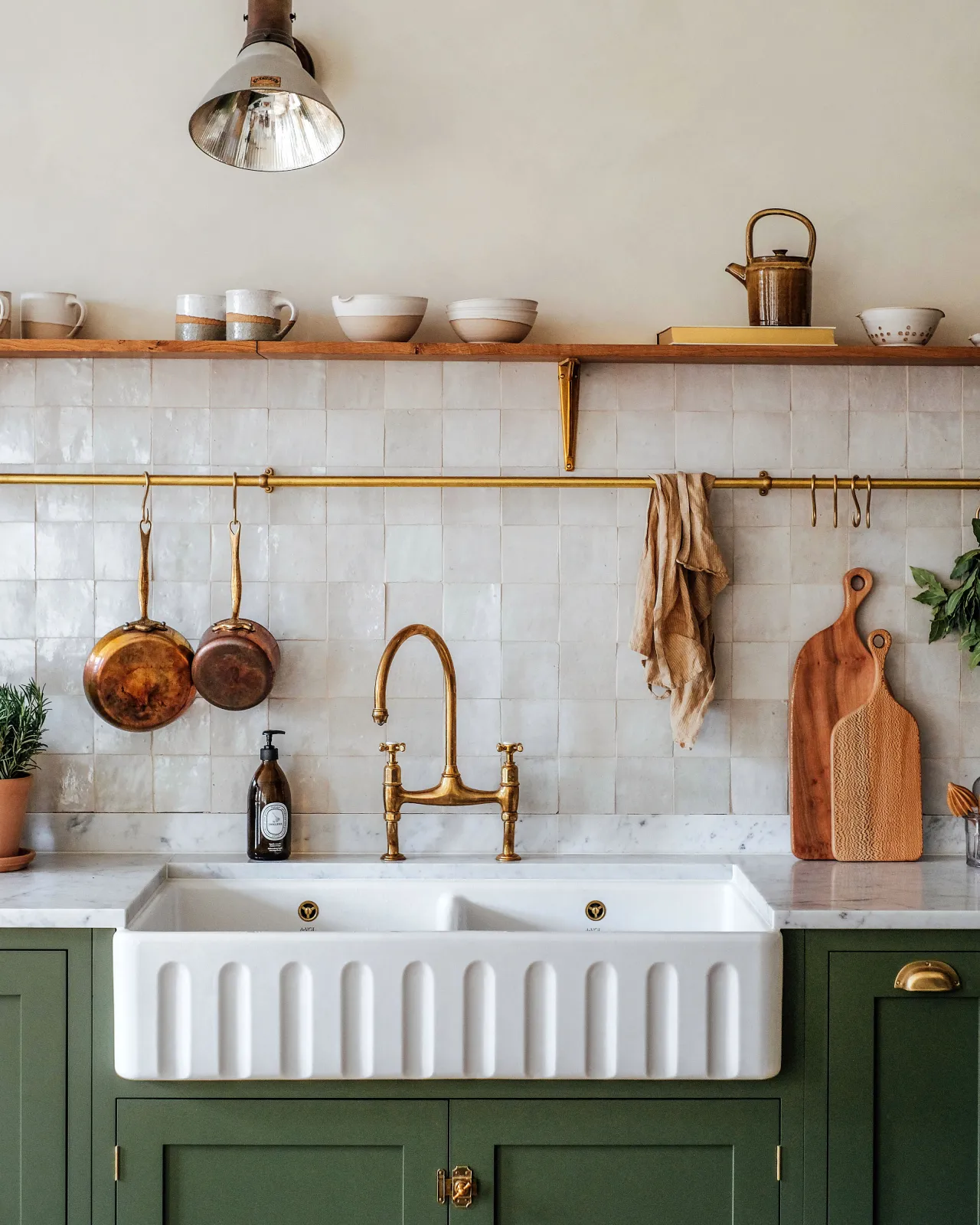 A straight-on view of a Shaker-style sink cupboard with a fluted Belfast sink, aged brass mixer tap and an aged brass hanging rail fixed to white metro tiles. Several vintage copper pots and linen tea towels hang via brass s hooks along the rail.