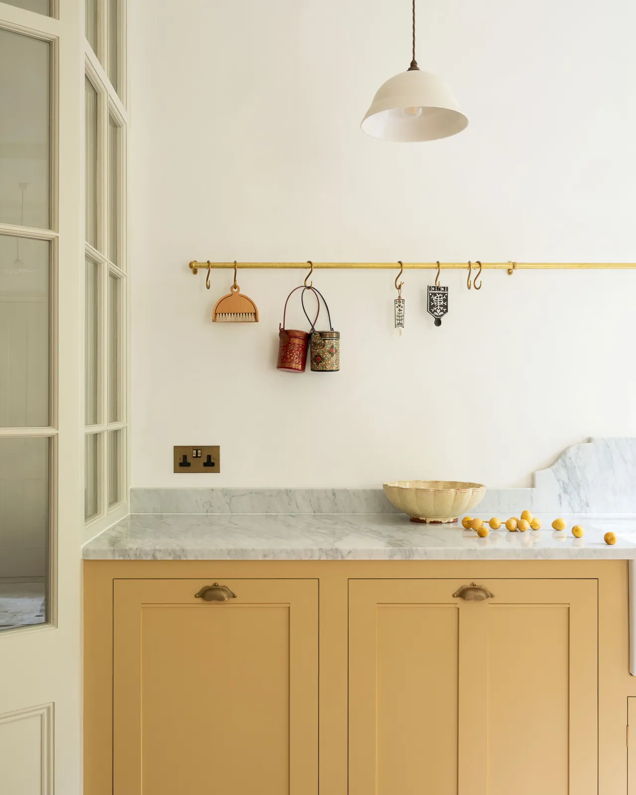 A close-up straight-on view of an aged brass hanging rail fixed to the wall above yellow painted Georgian-style kitchen cupboards. Several vintage utensils and decorations hang via brass s hooks along the rail.