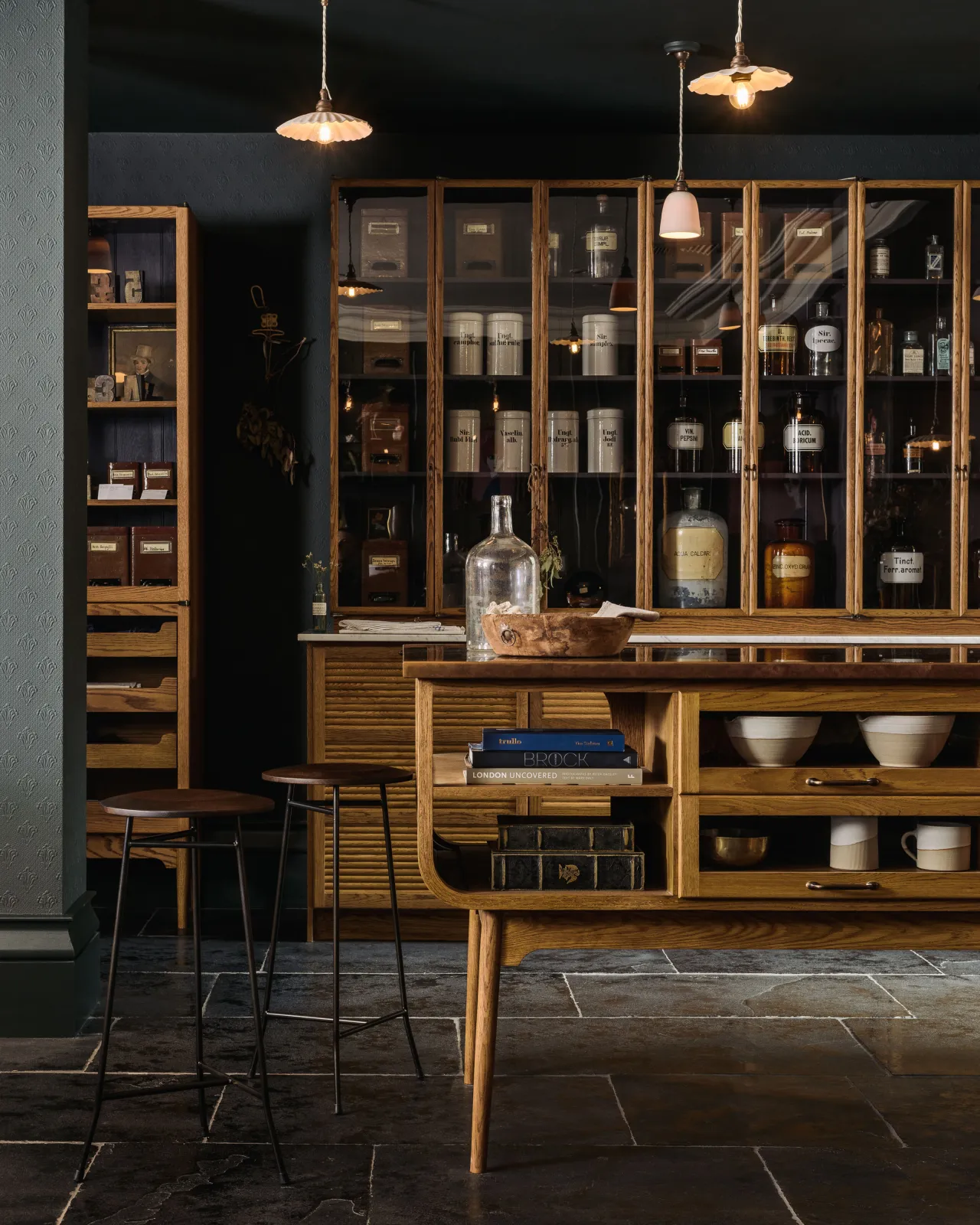 A view towards a bank of tambour-fronted and glazed Haberdasher's cupboards in deVOL's New York showroom, with dark painted walls and Brushed Charcoal Limestone flooring.