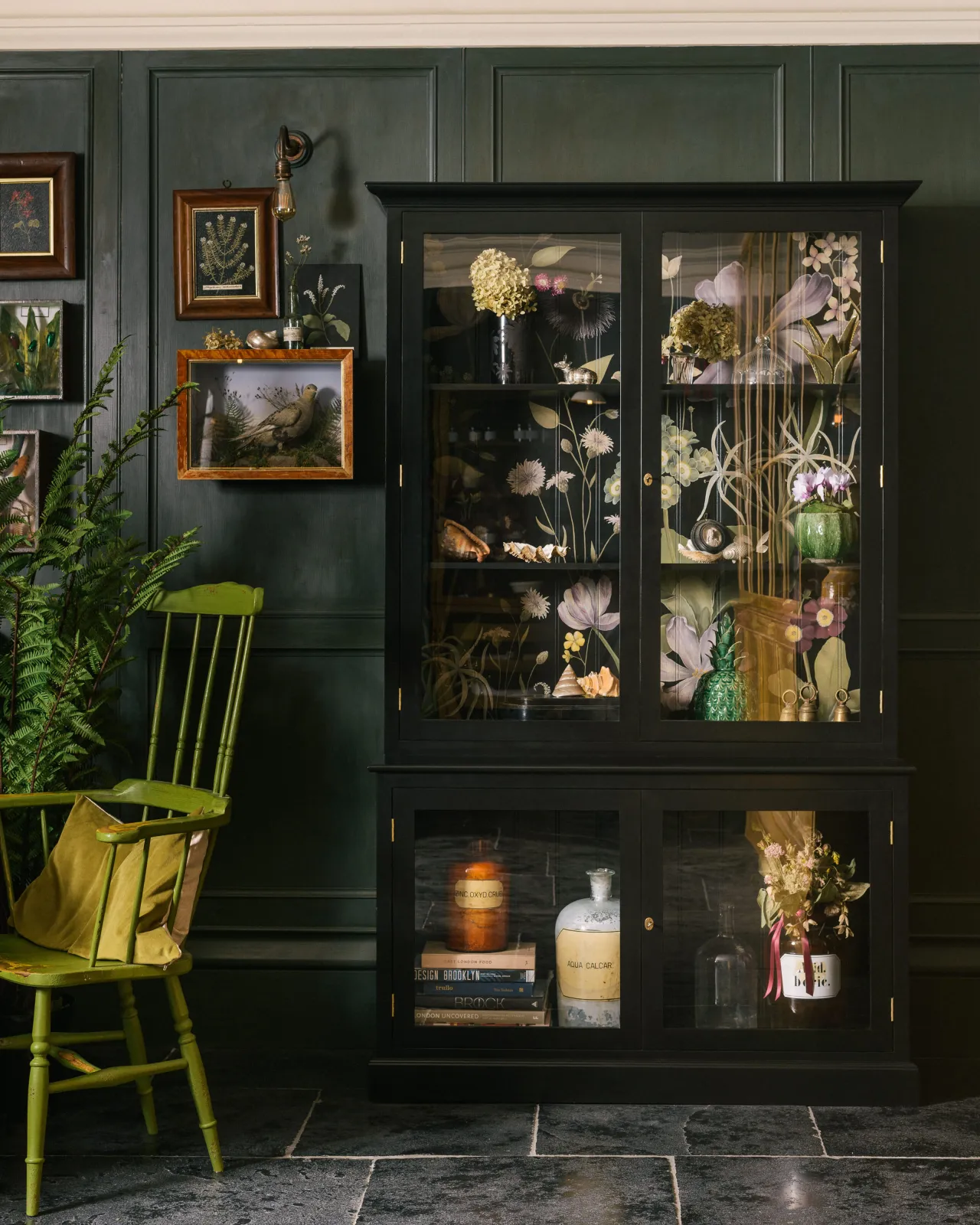 A straight-on shot of a black Curiosity Cupboard, situated in the dining area of deVOL's New York showroom. Dark green walls, Brushed Charcoal Limestone flooring and an antique chair, create the feeling of an aged, lived-in space.