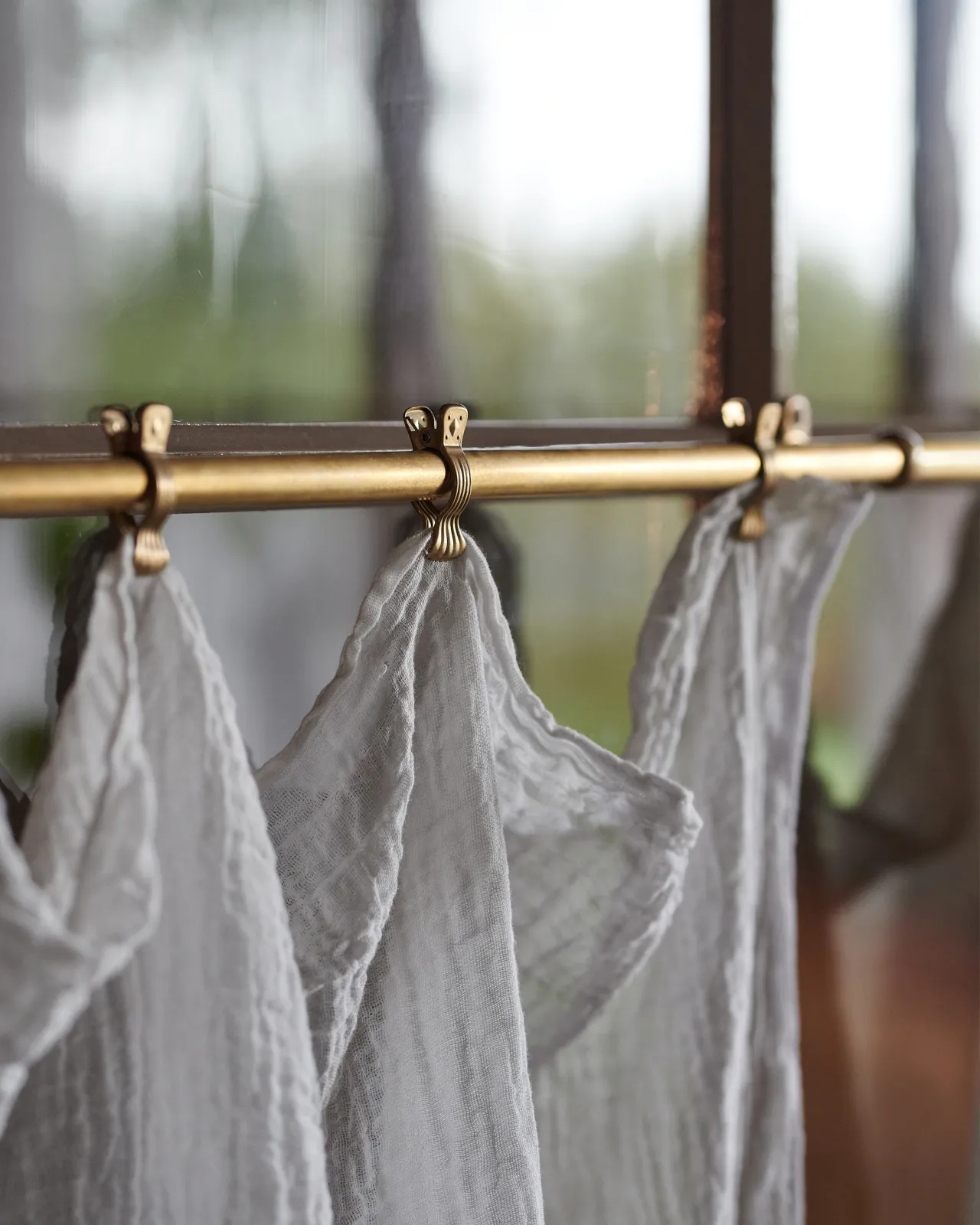 A close-up view of some small decorative brass crocodile-style clips on an aged brass curtain rail, holding a delicate linen curtain.