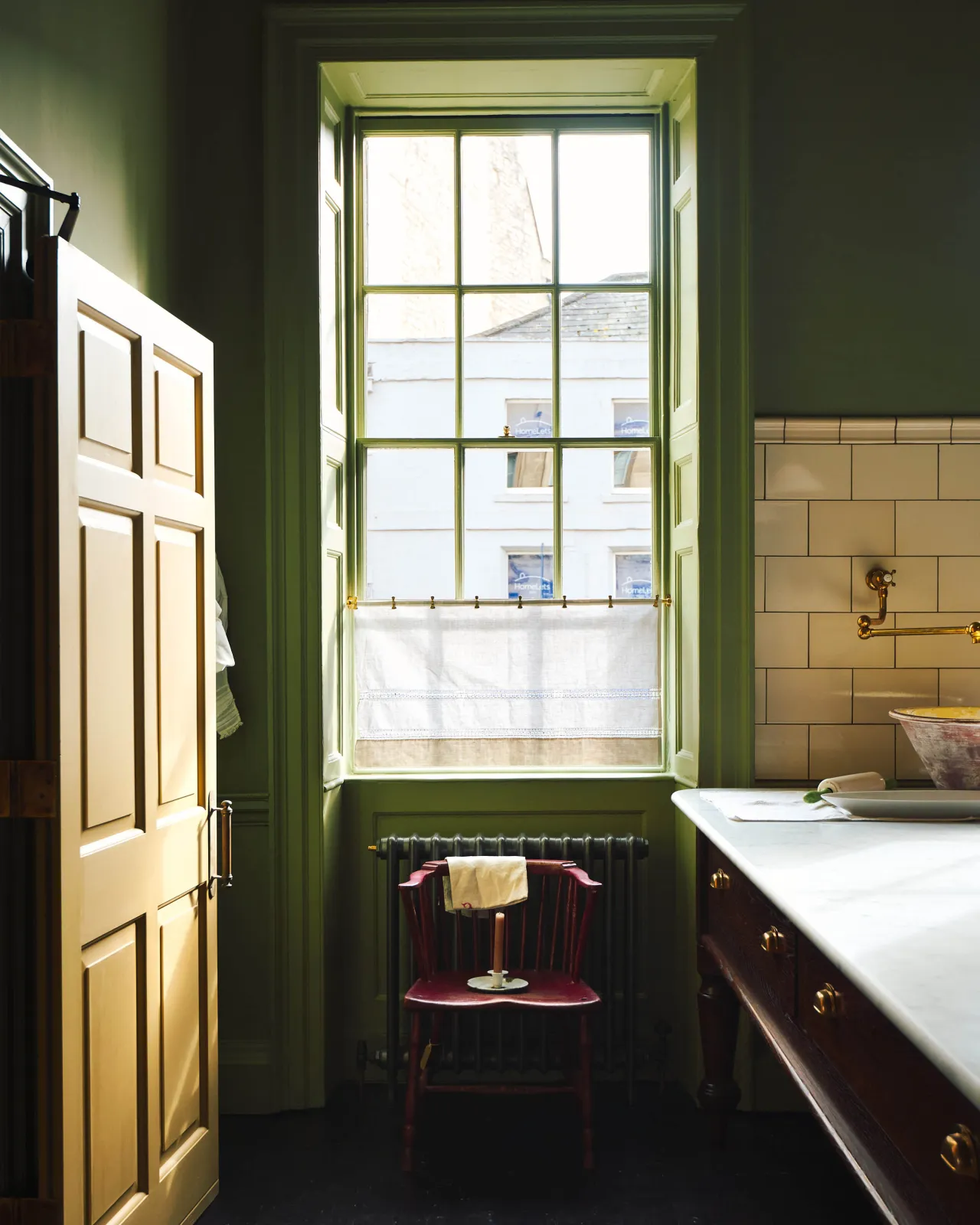 A straight-on view of a tall sash window in a Georgian interior with moody green painted walls and antique furniture. A short aged brass curtain rail holds a sheer linen curtain over the lowest window panes.