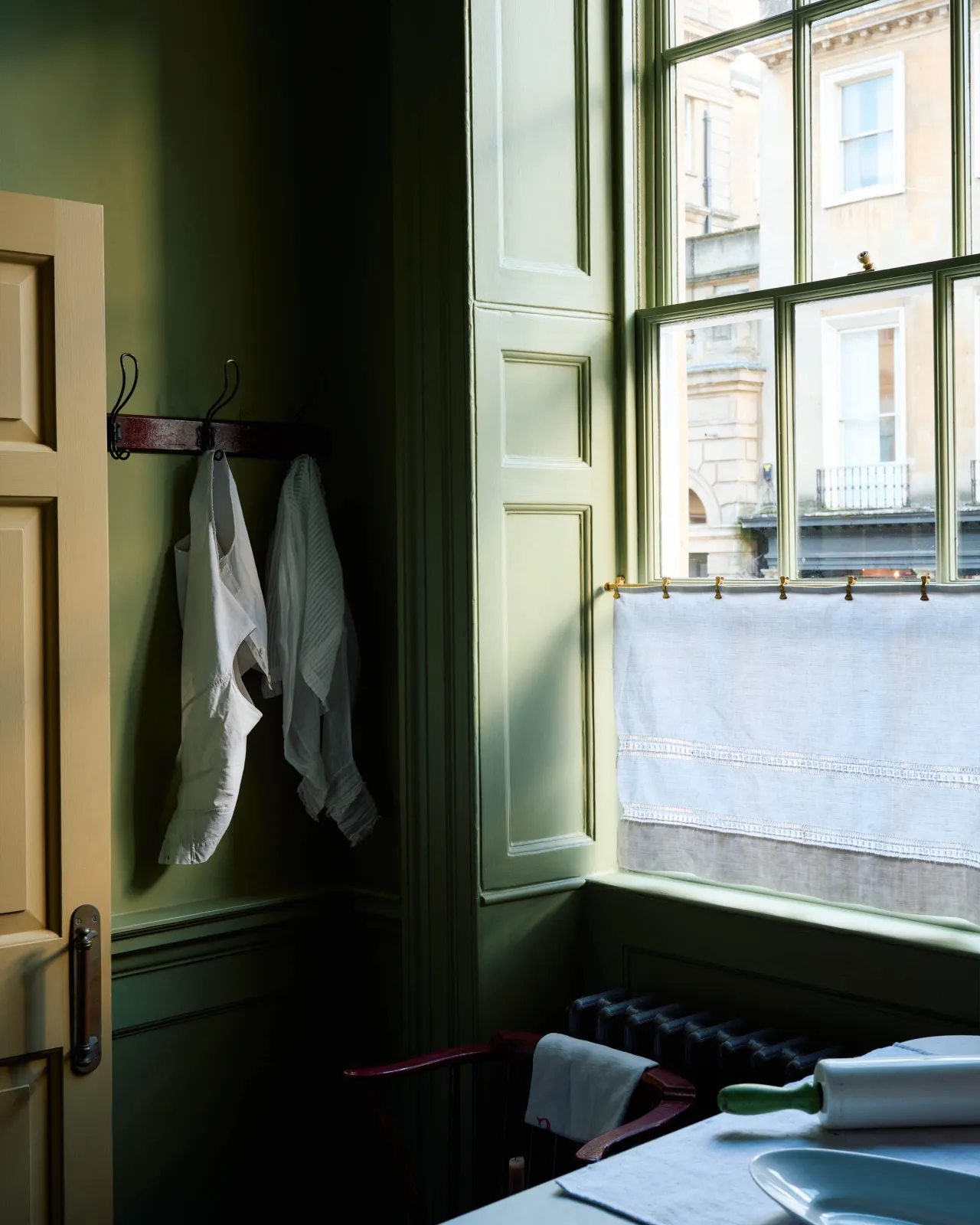 An oblique view of a short aged brass curtain rail hanging a sheer linen curtain in front of a tall sash window in a Georgian interior with moody green painted walls and original painted woodwork.