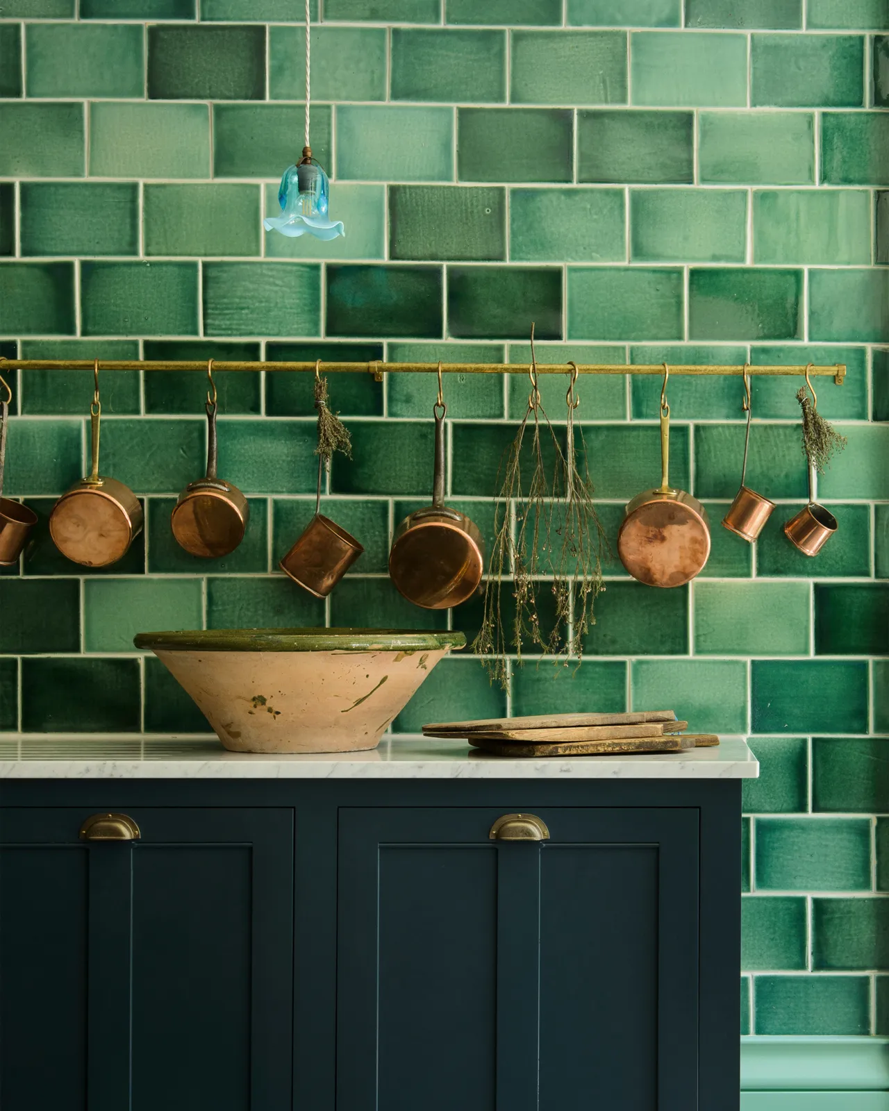 A large installation of handmade glossy emerald green wall tiles with royal blue painted Georgian-style kitchen cupboards, Carrara marble worktop and an aged brass hanging rail decorated with vintage copper pans.