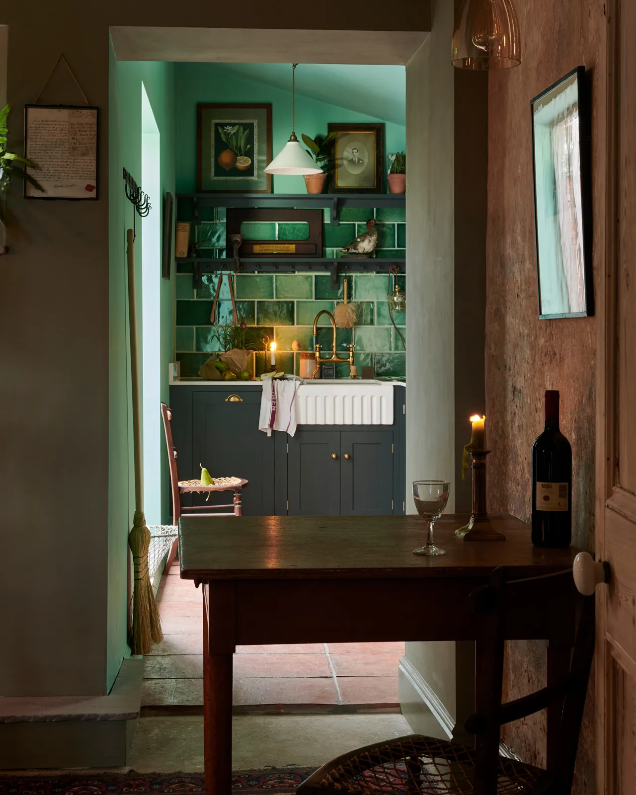 Looking through an open doorway into a small Shaker-style kitchen with dark green painted cupboards and a large wall of glossy emerald green wall tiles. A lit candle flickers on a table in the foreground.