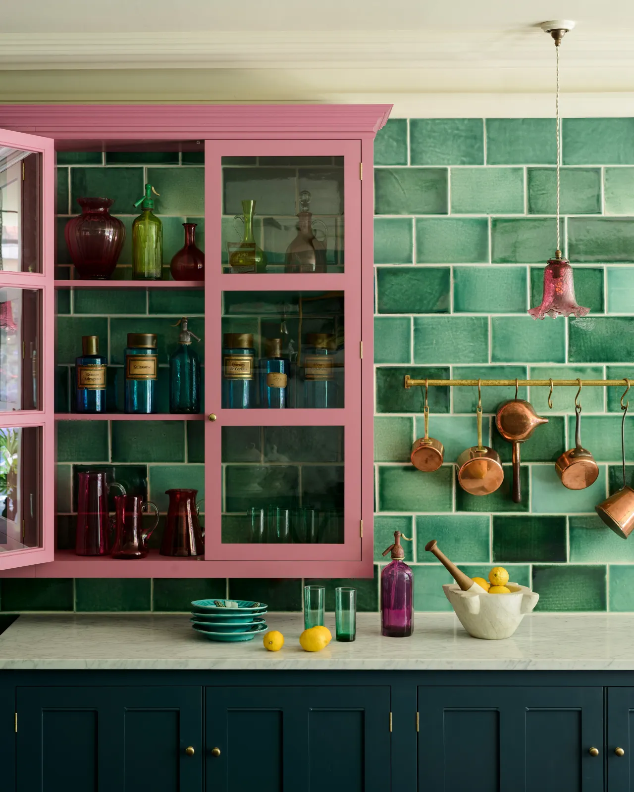 A straight-on image of some glossy emerald green wall tiles fitted behind a vibrant flamingo-pink glazed wall cupboard in deVOL's Manhattan showroom. Copper accents, colourful glassware, and a pop of yellow from scattered lemons complete the striking vignette.
