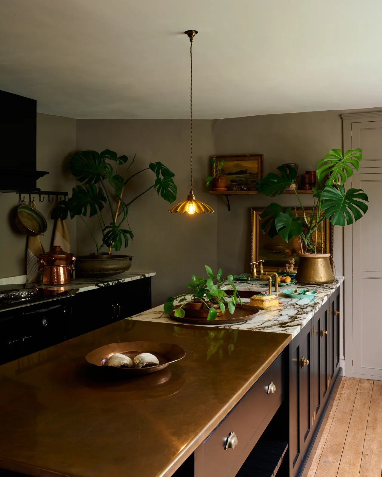 An oblique view of an atmospheric kitchen with black painted Shaker-style cabinetry, grey walls, wooden floorboards, vintage styling and large monstera plants. A small frilly chartreuse glass pendant light with brass fittings and brown twisted fabric flex cord is hanging above an aged copper worktop.