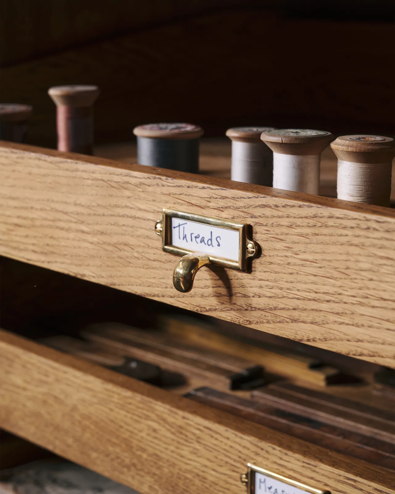 A close-up oblique view of a small cardframe pull handle in a warm burnished brass finish and a handwritten paper label that reads 'Threads', fitted to an oak drawer front partially concealing spools of coloured thread.