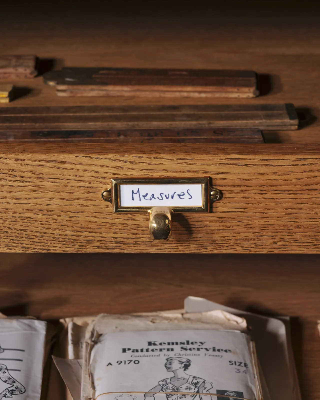 A close-up straight-on view of a small cardframe pull handle in a warm burnished brass finish and a handwritten paper label that reads 'Measures', fitted to an oak drawer front.