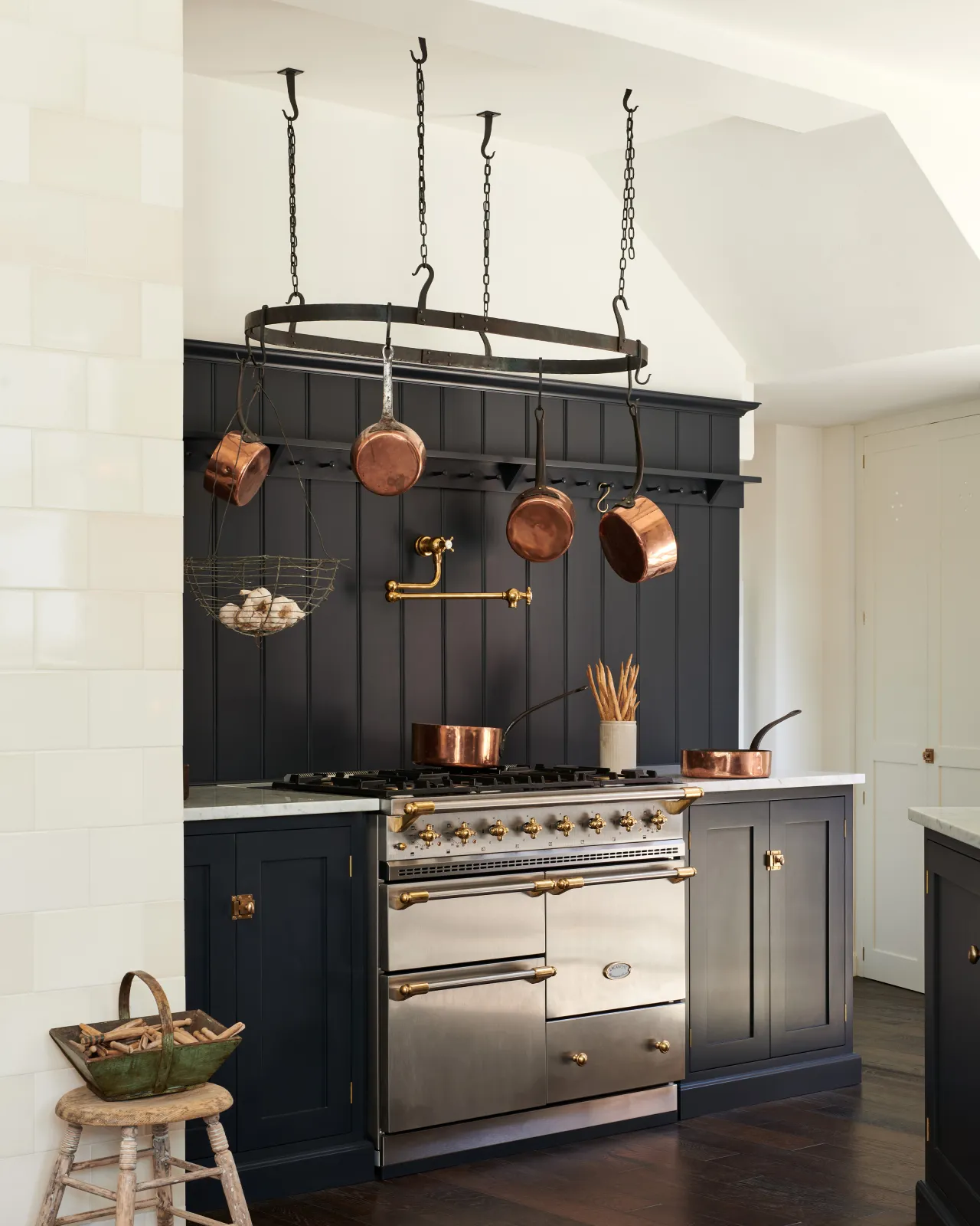 An oblique view of a simple Shaker-style kitchen with dark blue painted cabinetry and a stainless steel Lacanche range cooker with a large oval-shaped wrought iron pot hanger suspended from chains above.