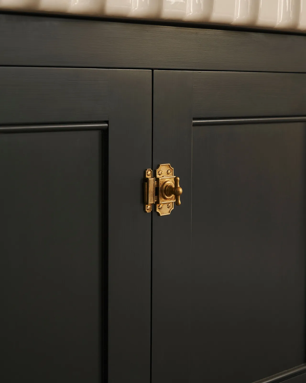 A close-up oblique view of a Victorian-inspired rack and pinion cupboard catch in a warm burnished brass finish, fitted to a black painted Georgian-style kitchen cabinet.