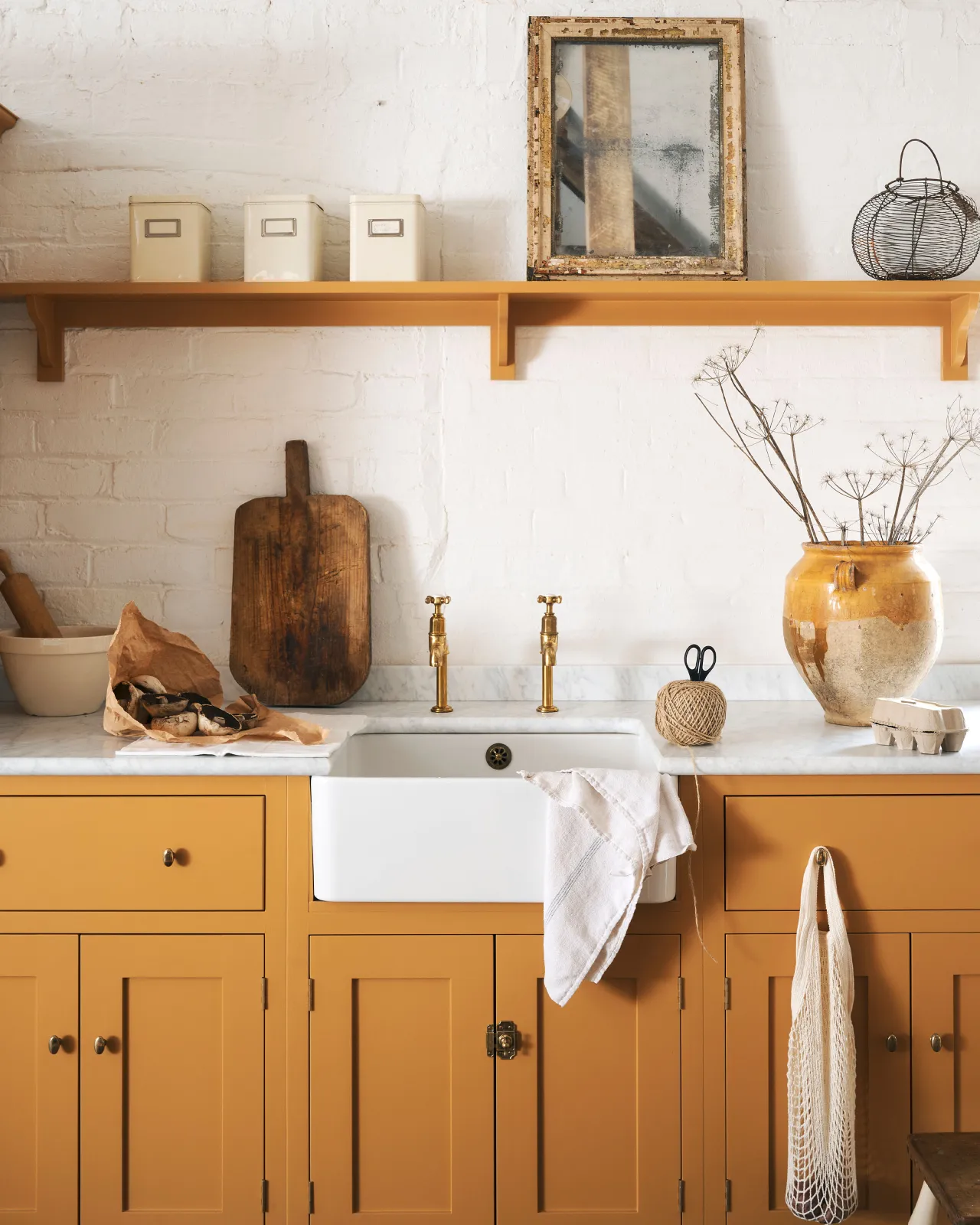 A straight-on view of a Victorian-inspired rack and pinion cupboard catch in a warm burnished brass finish, fitted to a yellow painted Shaker-style kitchen sink cabinet with a Belfast sink and Carrara marble worktop.