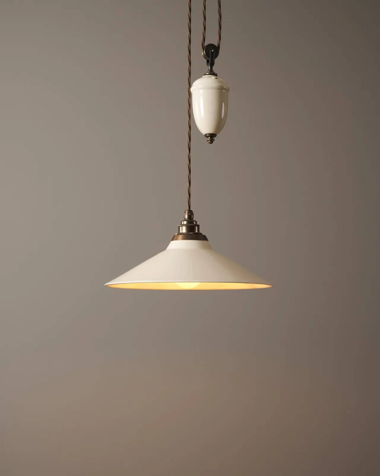 A rise and fall light with a small cream glazed ceramic shade and counterweight, an aged brass E27 lampholder and brown twisted fabric flex cord, the light is turned on.