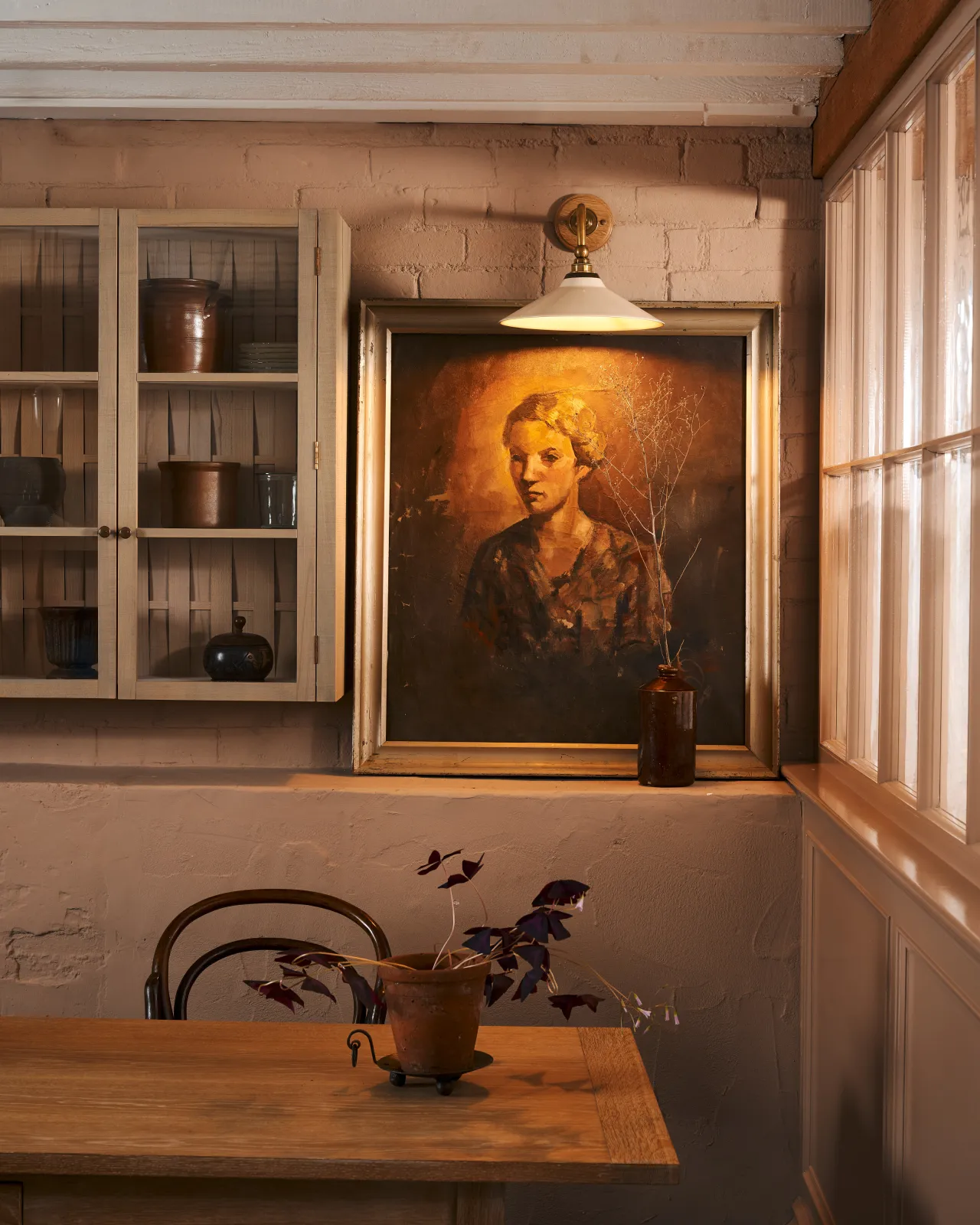 A straight-on view of a small Victorian-inspired wall light with a brass arm and a cream glazed ceramic shade fixed to a pink brick wall, beside a glazed wall cupboard, above an antique portrait painting.