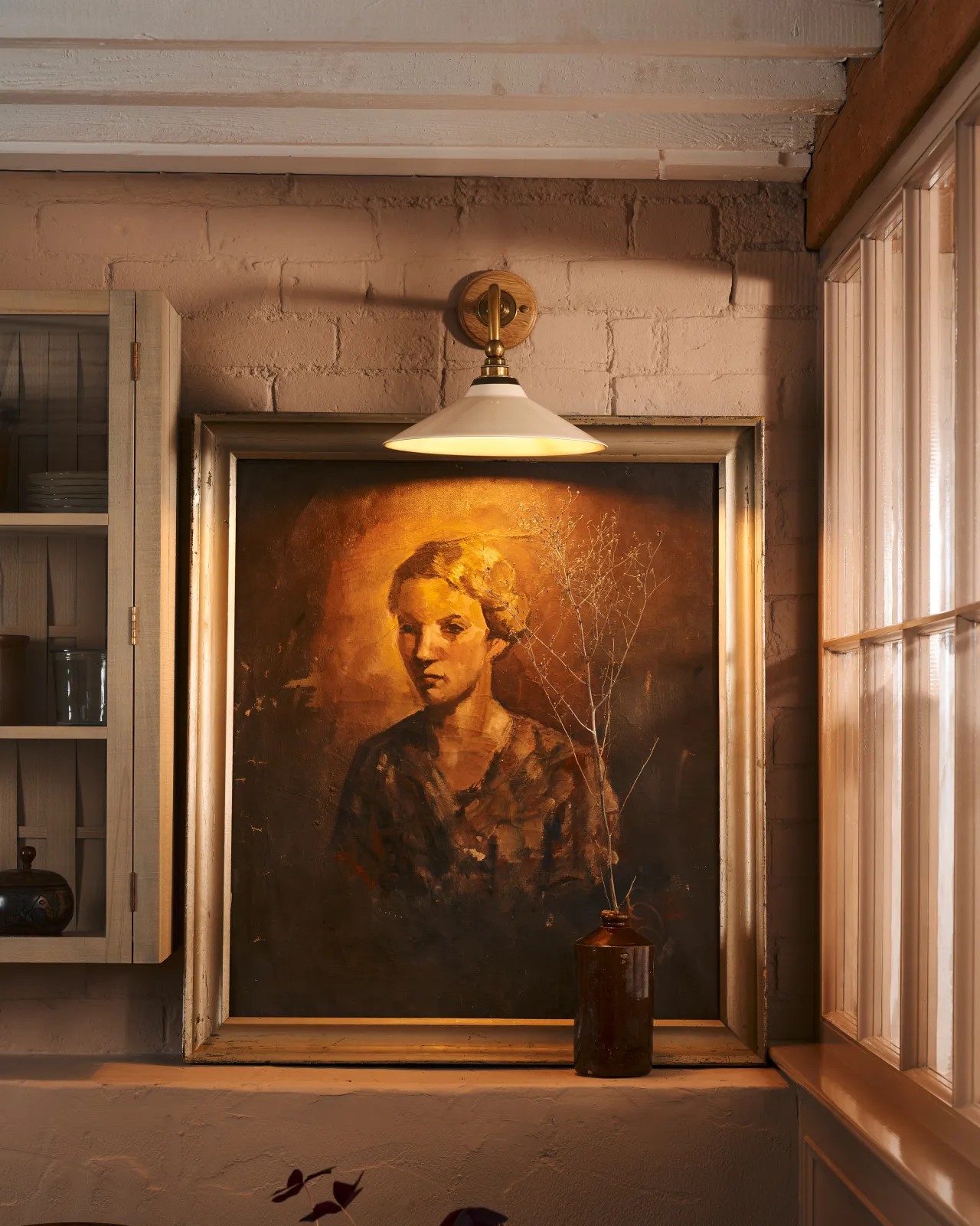 A close-up straight-on view of a small Victorian-inspired wall light with a brass arm and a cream glazed ceramic shade fixed to a pink brick wall above an antique portrait painting.