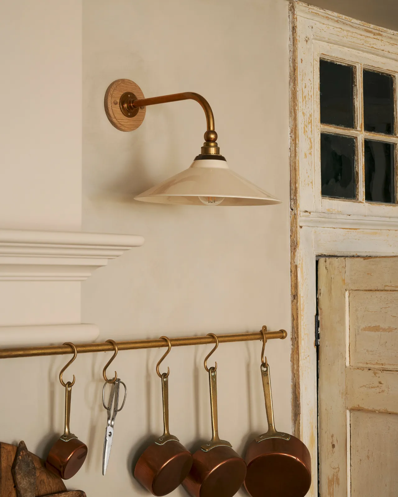 A close-up oblique view of a small Victorian-inspired wall light with a brass arm and a cream glazed ceramic shade fixed to a cream wall above an aged brass hanging rail.