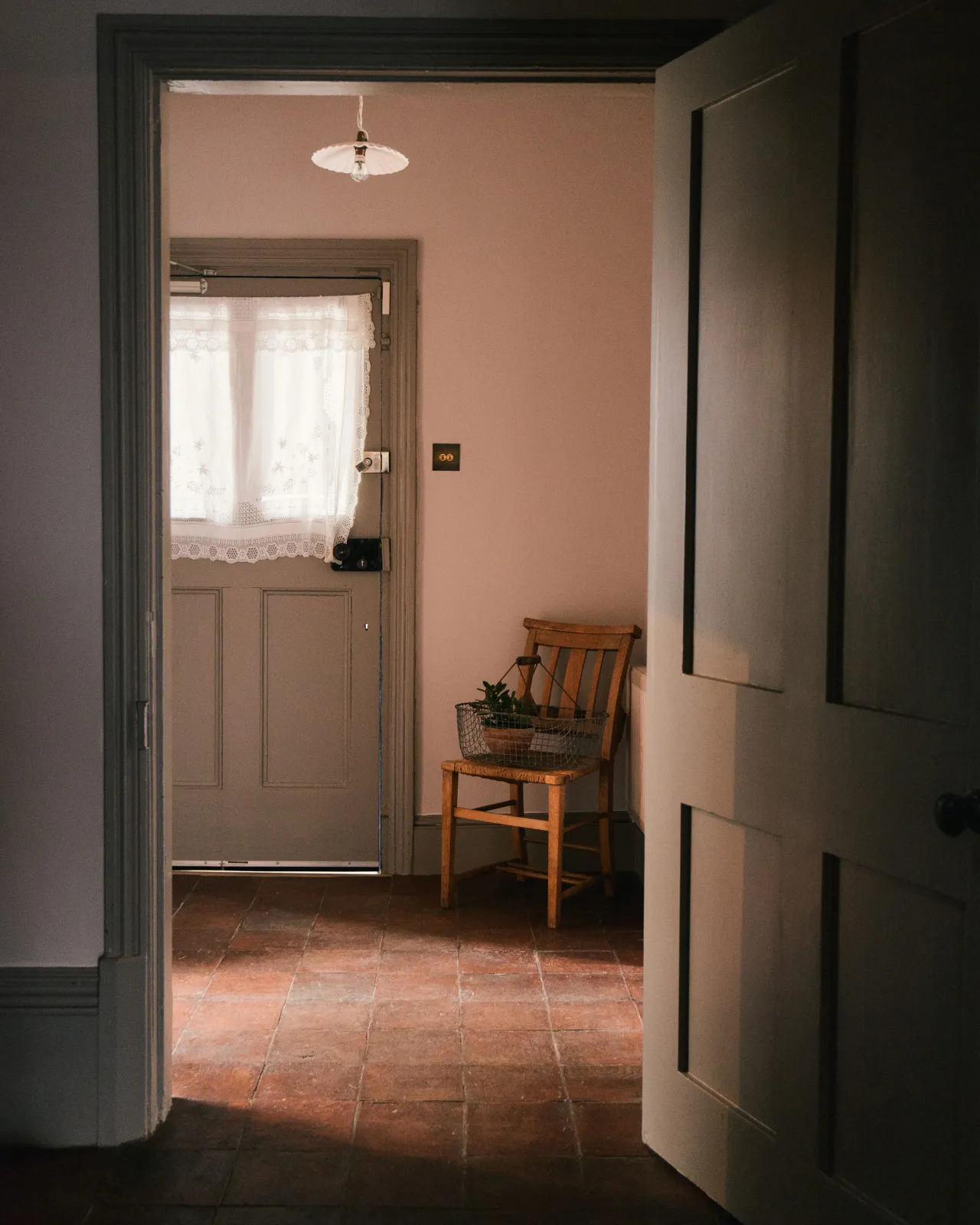 Looking through a cottage doorway with white walls, grey painted woodwork and an antique wooden chair on square terracotta flooring with an authentic aged finish.