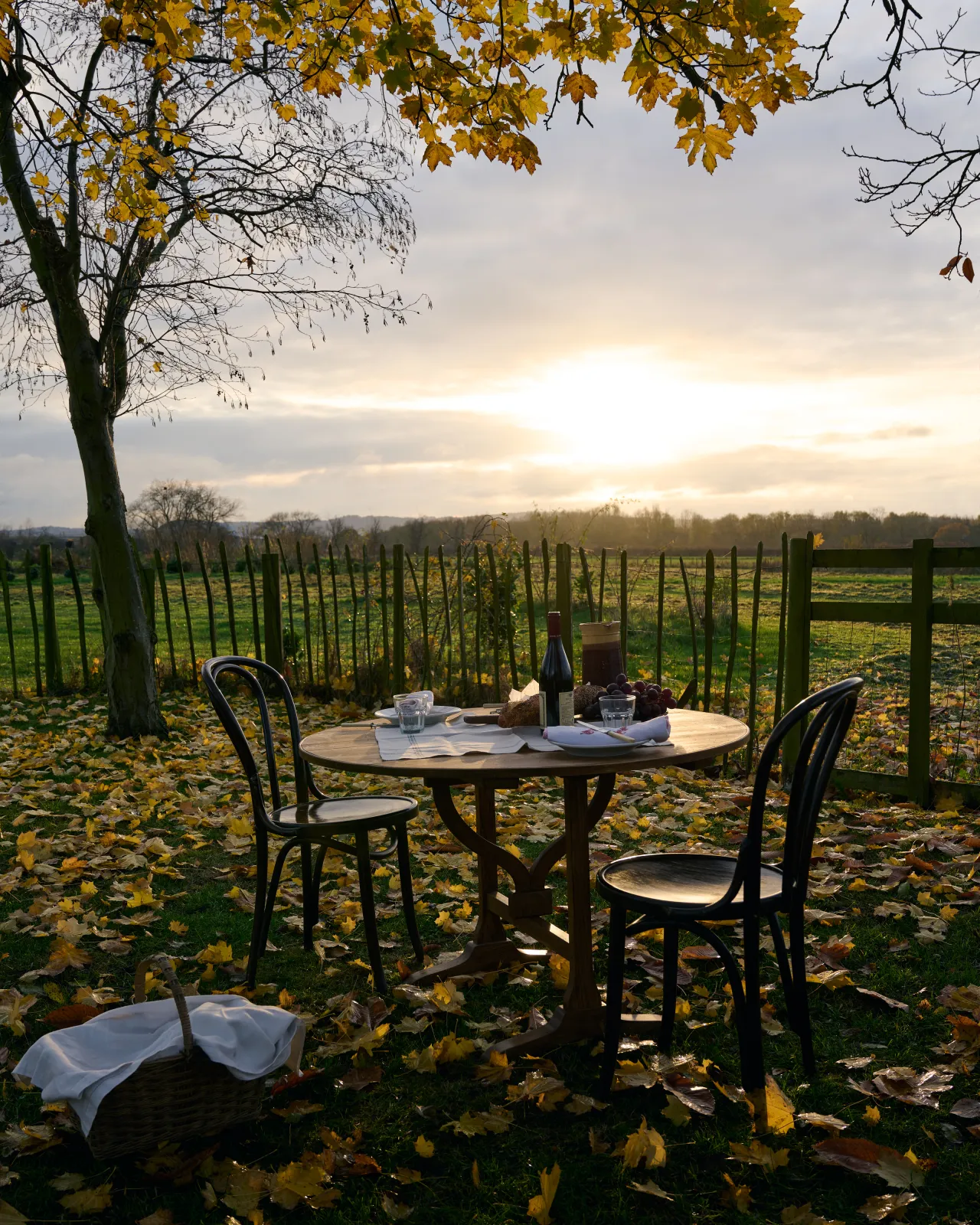 A round pine dining table with vintage wooden chairs outdoors under trees with browning leaves and an autumn sunset in the distance.