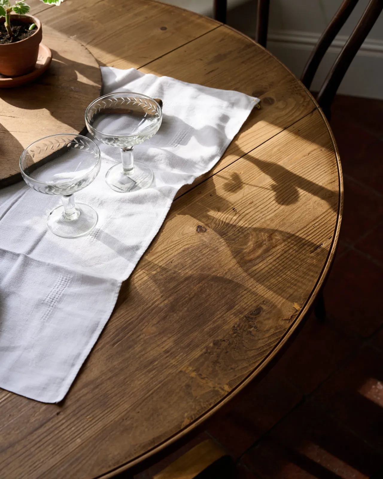 A close-up view of a round pine dining table with a small vintage linen tablecloth and clear wine glasses.
