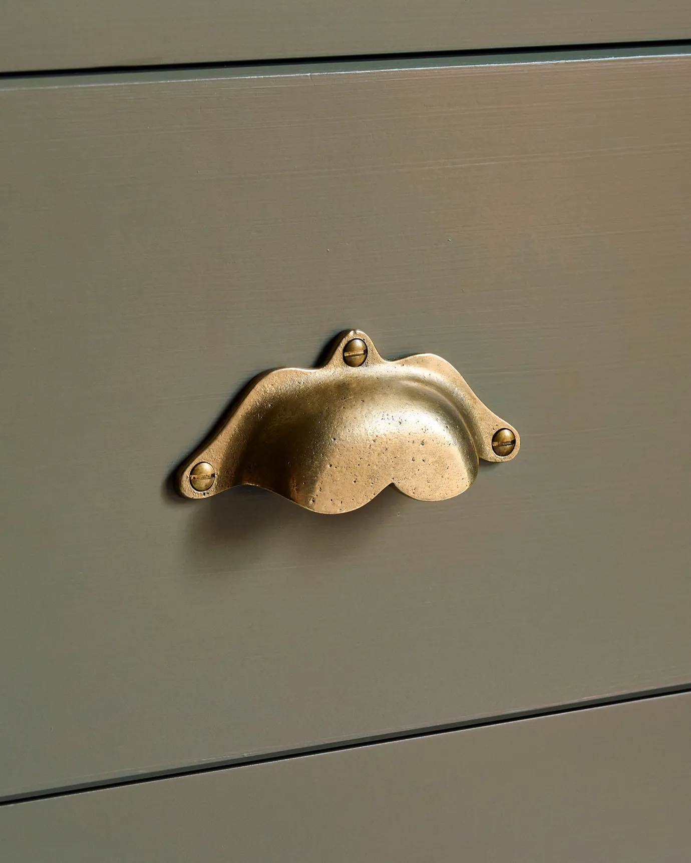 A close-up oblique view of a bohemian-style cup handle in an aged brass finish fitted to a kitchen cupboard drawer painted in a moody sage green colour.