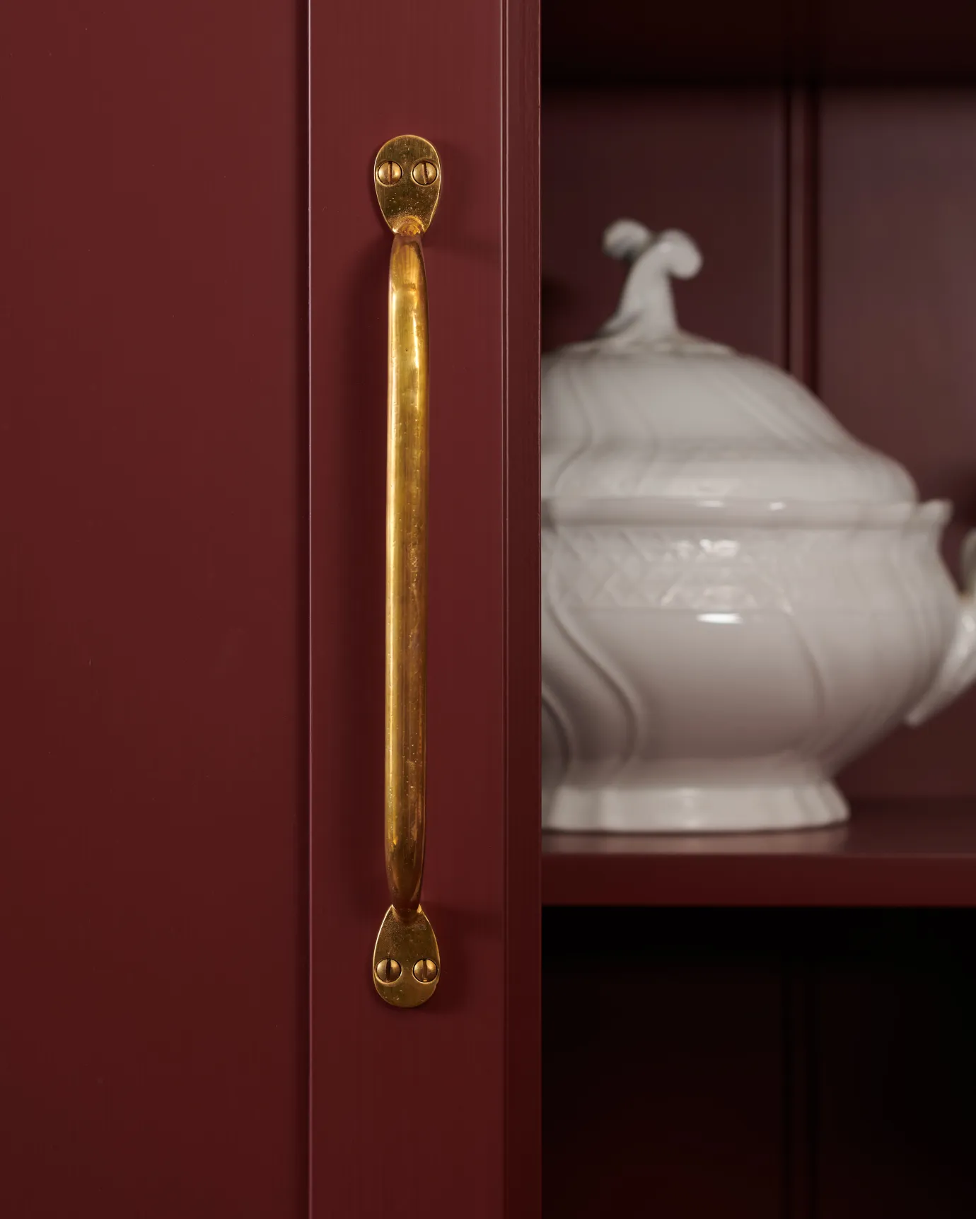 A close-up straight-on view of a classic pull handle in a warm burnished brass finish fitted to a wine-red coloured pantry door. Inside the cupboard, there is an ornate vintage white dish.