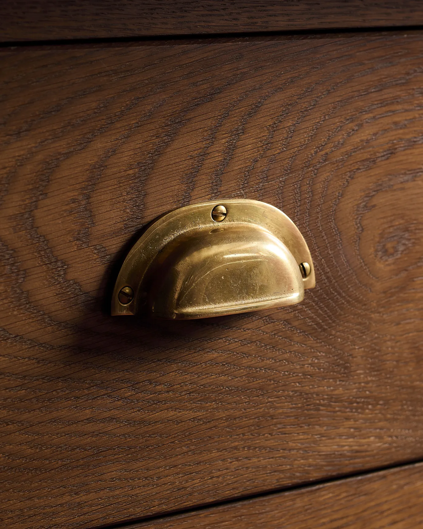 A close-up oblique view of a classic cup handle in a warm burnished brass finish fitted to a kitchen cupboard drawer with a dark stained oak finish.