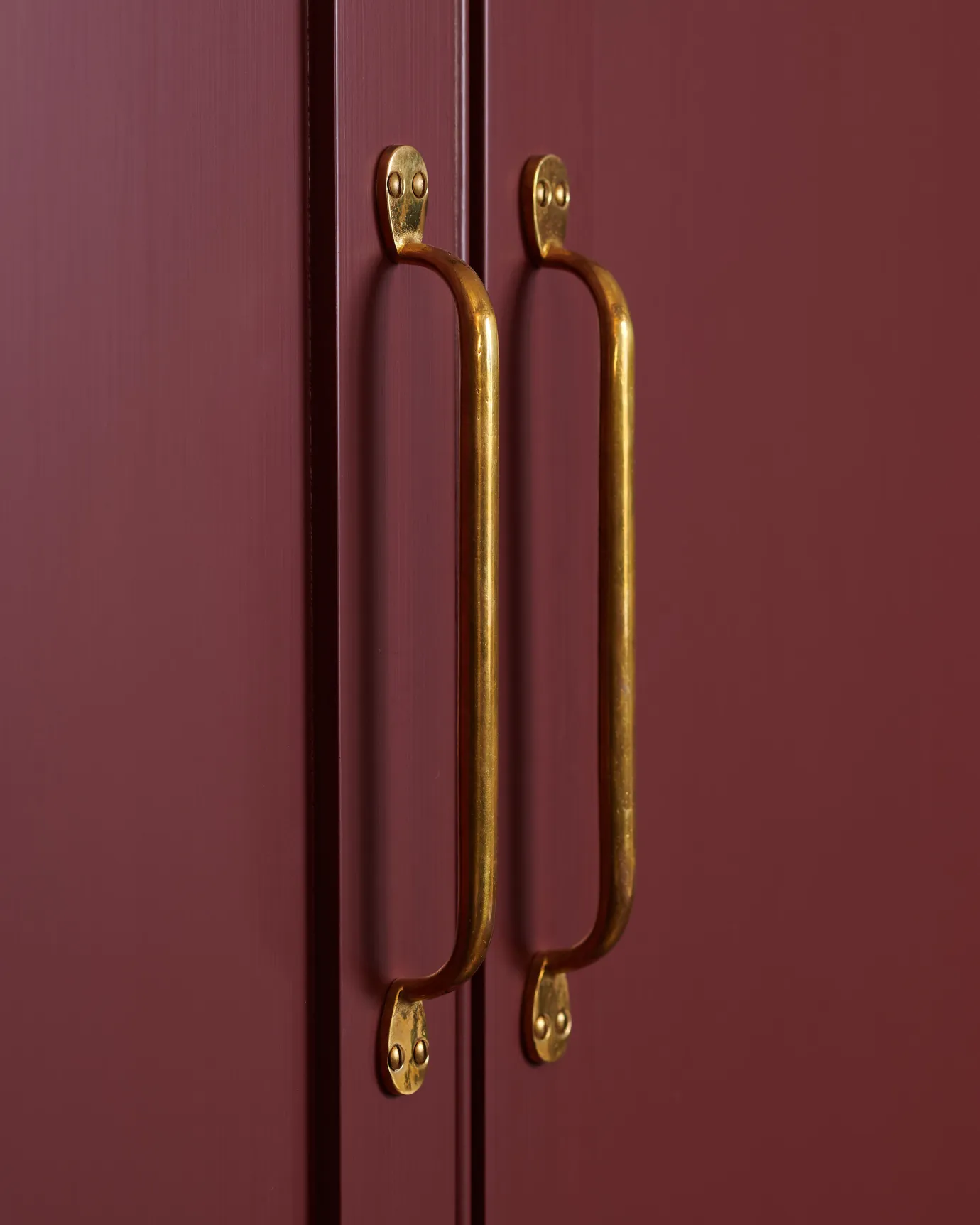 A close-up oblique view of a pair of classic pull handles in a warm burnished brass finish fitted to pantry doors painted in a wine-red colour.