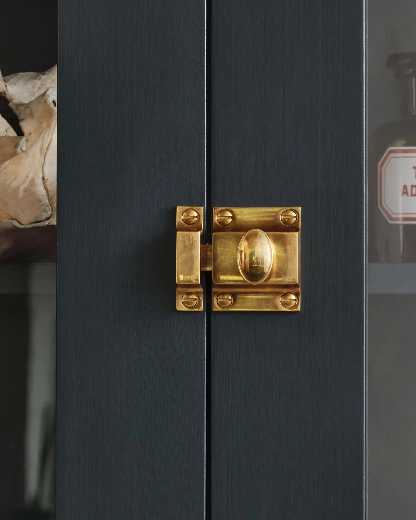 A straight-on view of a classic-style square cupboard catch with an ornate oval-shaped knob in a warm burnished brass finish fitted to dark blue painted glazed cupboard doors.