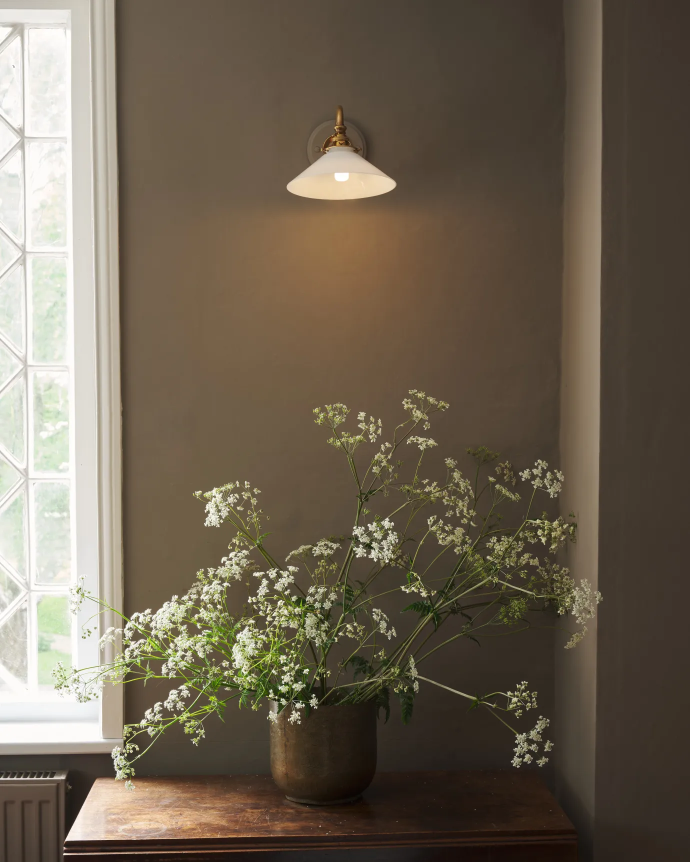 A straight on view of an elegant wall light with a curved brass arm, painted wooden pattress and small milk glass shade, fitted to a grey wall beside a leaded window and above large sprigs of cow parsley. The light is on and casting a warm glow over the wall below.