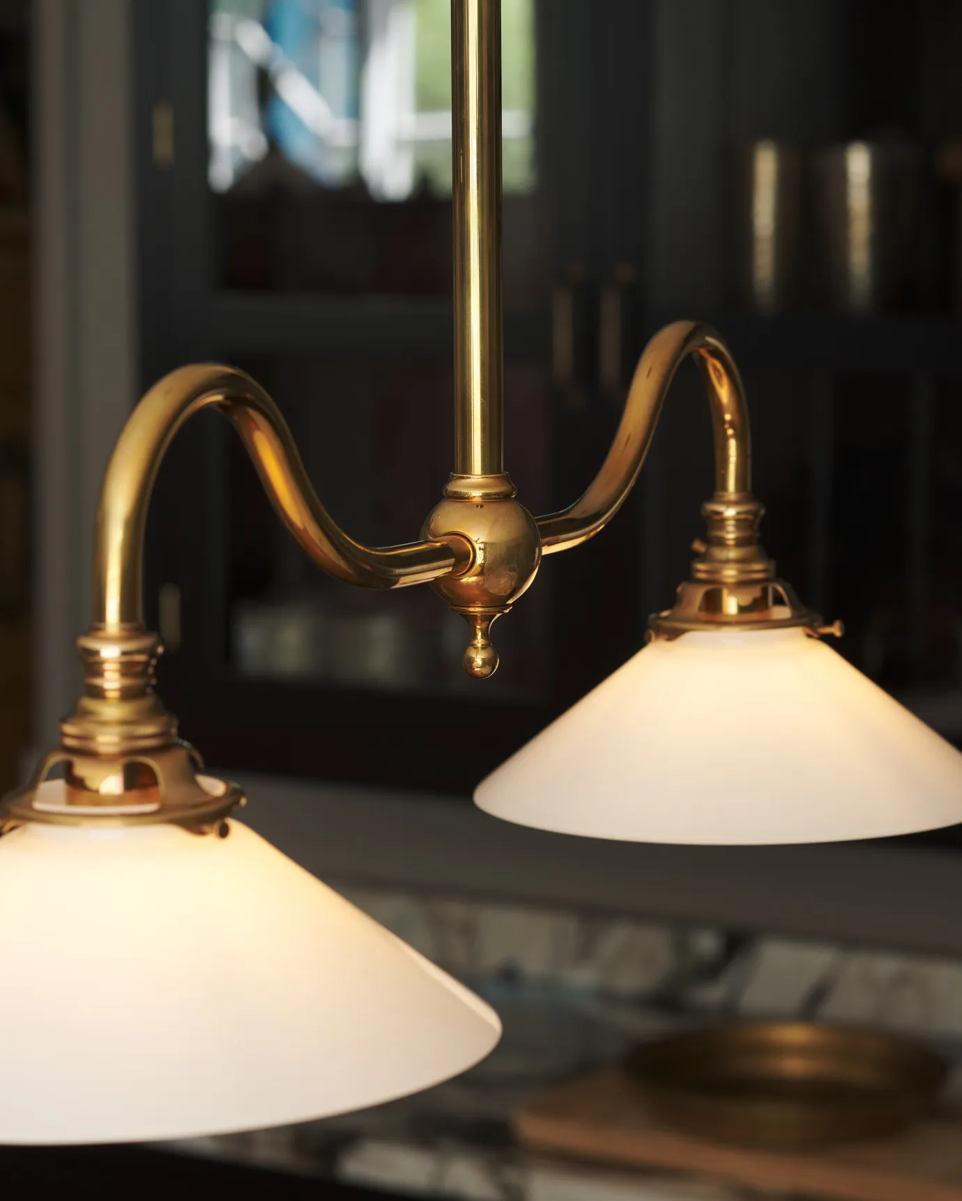 Two small milk glass light shades connected to two curved brass arms, extending from a central ball joint with a decorative finial and central pole.