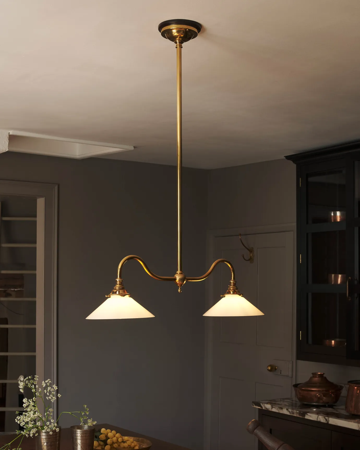 An elegant ceiling light with a rigid brass pole and two curved arms, each holding an illuminated small milk glass shade. The light gives off a warm glow above an electric and atmospheric dark kitchen.
