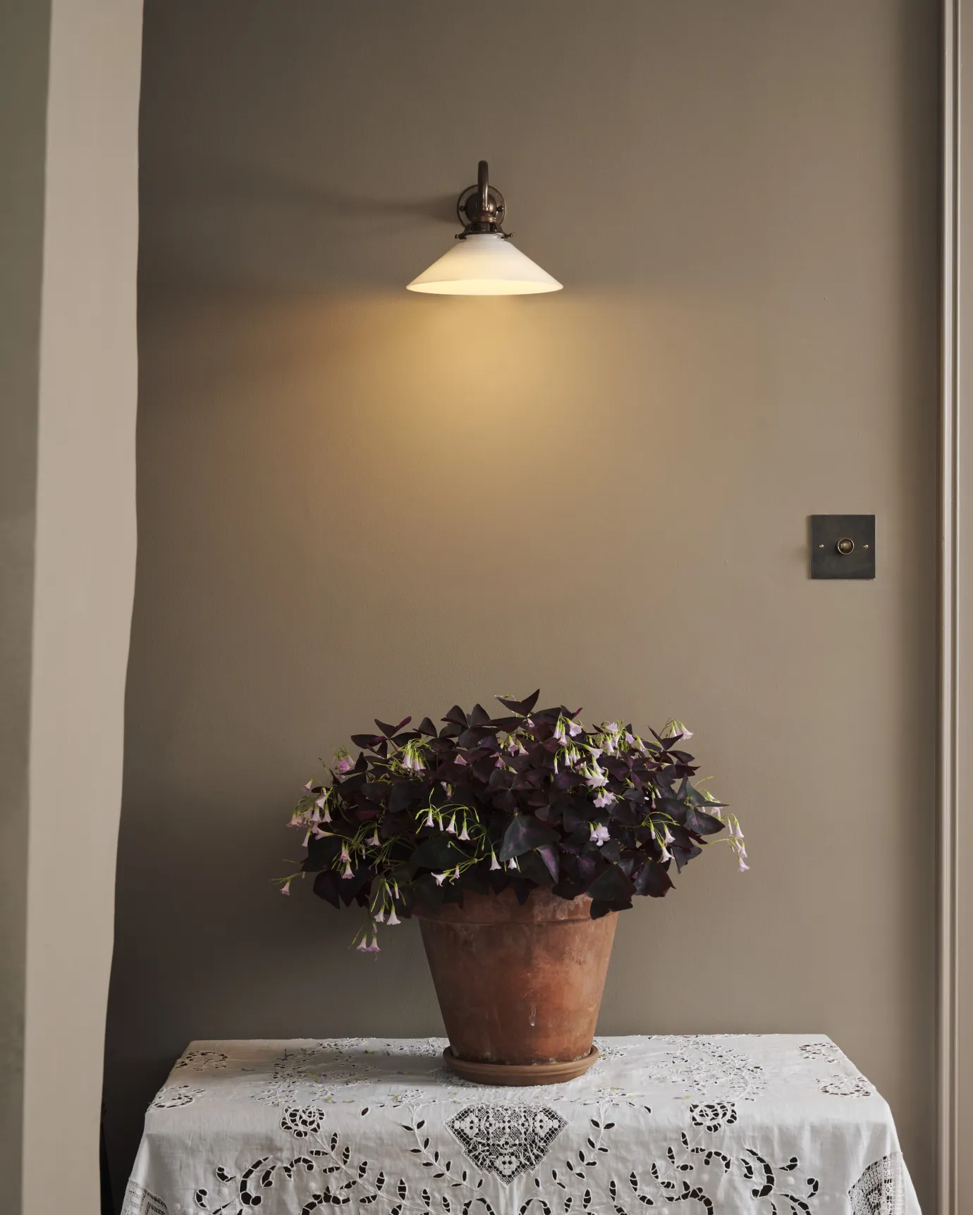 A small milk glass wall light above a large potted plant on a lace tablecloth. The light is on and casting a warm glow across the wall.