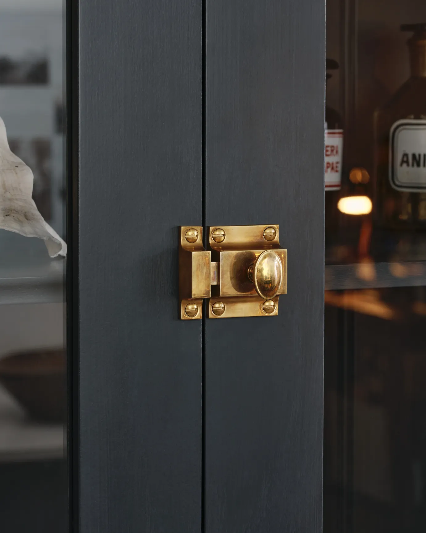 An oblique view of a classic-style square cupboard catch with an ornate oval-shaped knob in a warm burnished brass finish fitted to dark blue painted glazed cupboard doors.
