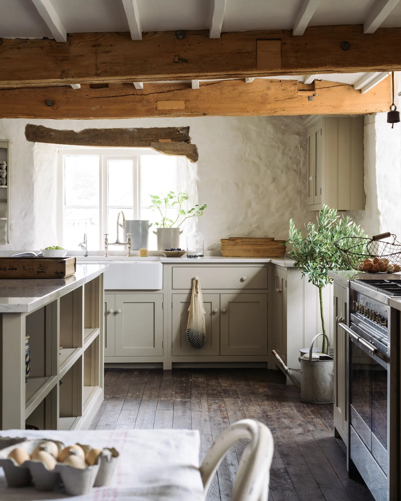 A large cottage kitchen with creamy mushroom coloured Shaker-style kitchen cupboards, dark worn floorboards and large wooden ceiling beams.