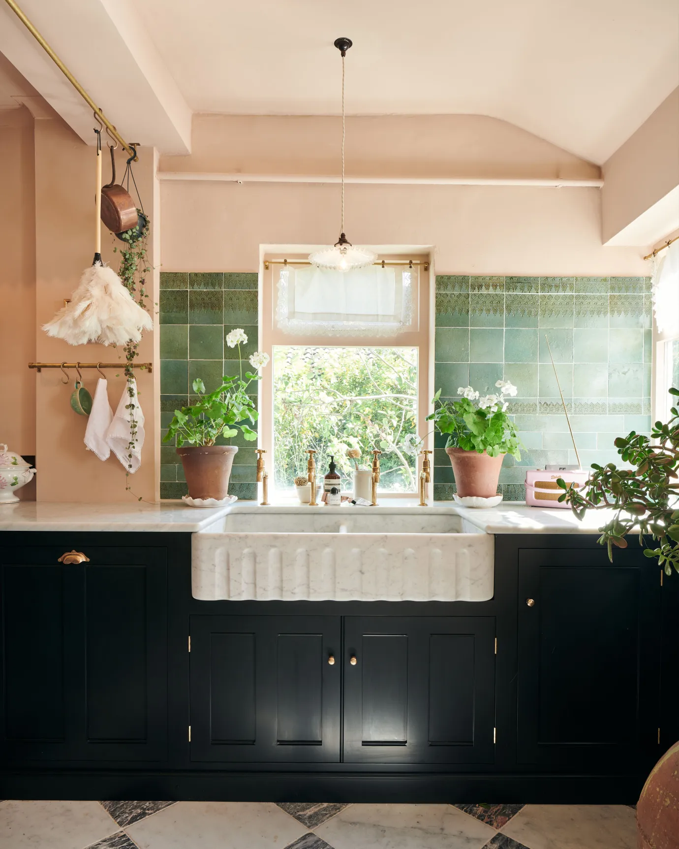 A straight-on view of a black painted Georgian-style kitchen sink cabinet with a large fluted Carrara marble sink, two sets of aged brass bibcock taps, pink walls and teal patterned wall tiles.