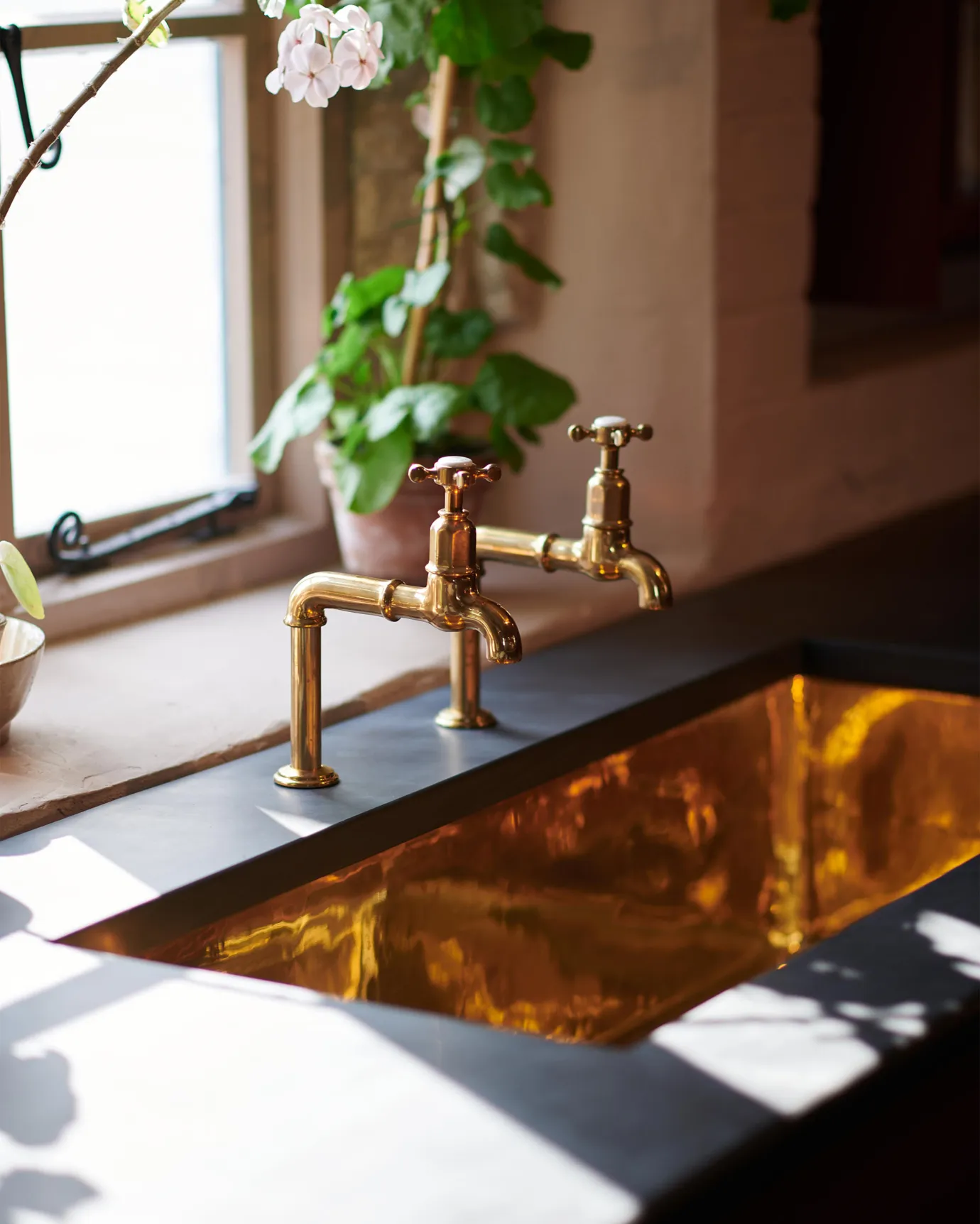 An oblique view of a pair of traditional-style aged brass bibcock taps above a grand polished brass sink and an understated slate worktop.
