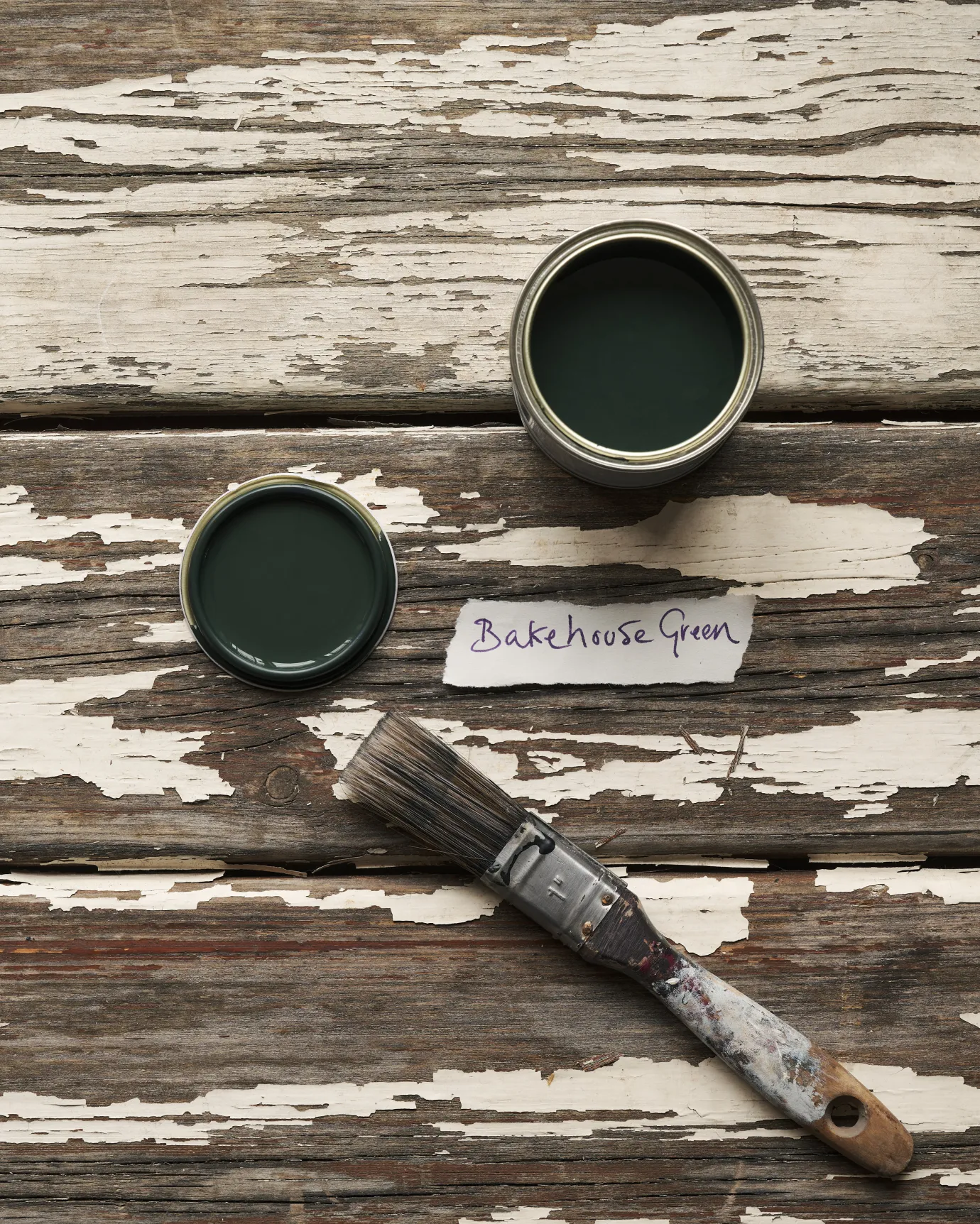 An overhead view of an open paint pot and a worn brush on a rustic tabletop. The dark green colour inside is labelled 'Bakehouse Green'.