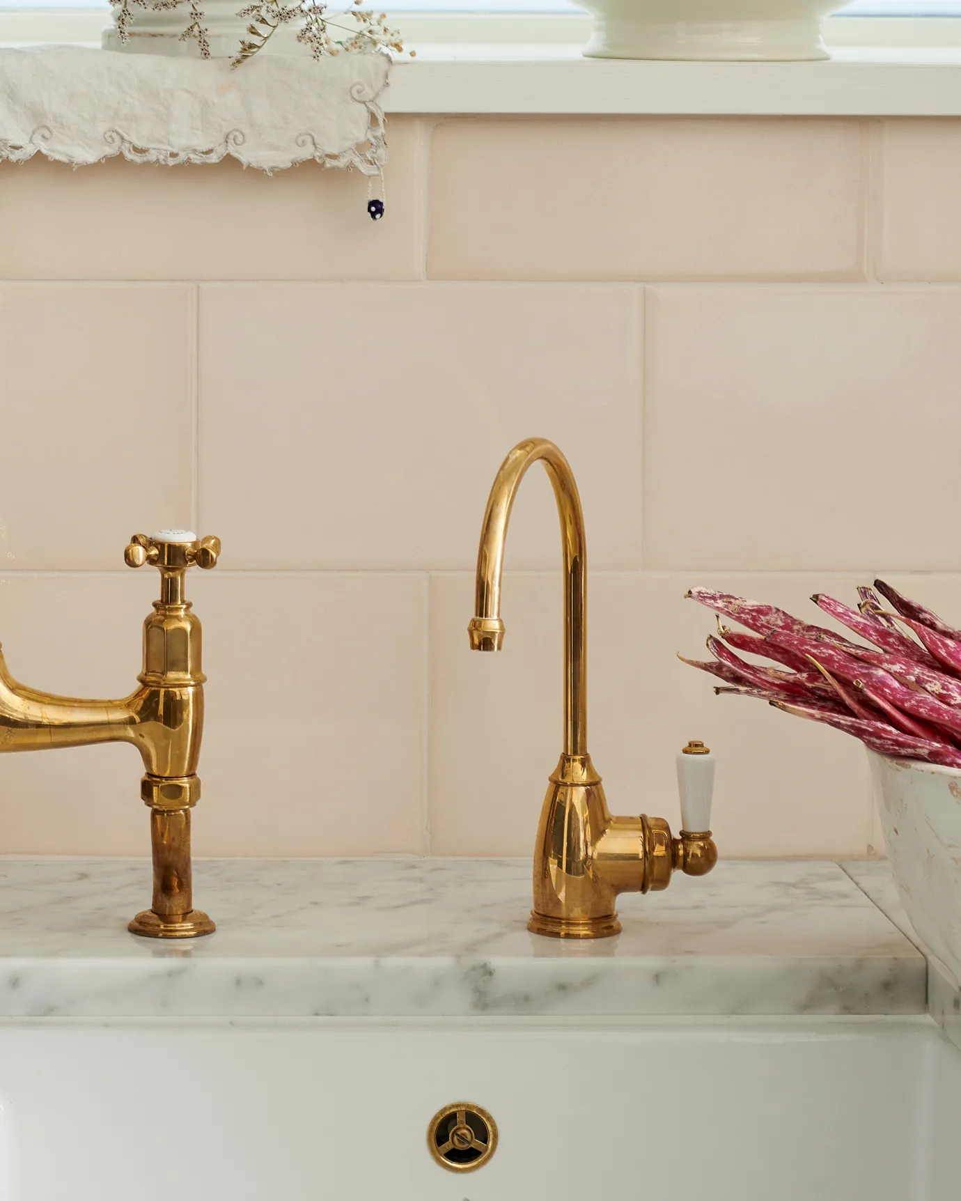 A straight-on view of a small traditional-style aged brass tap with a white ceramic lever with cream subway wall tiles behind.