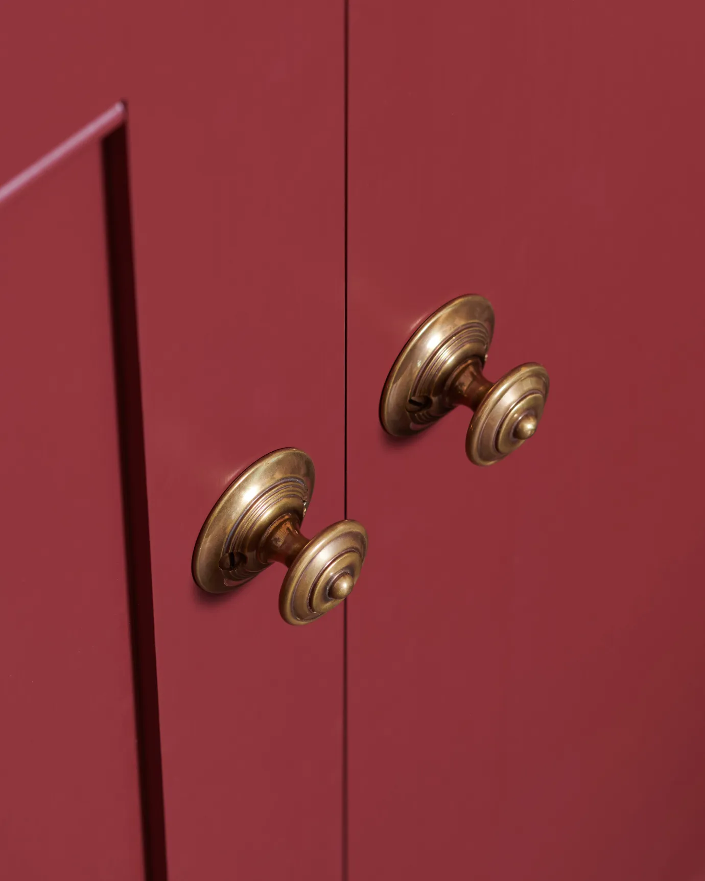 A close-up oblique view of a pair of Victorian-inspired ornate doorknobs in a warm burnished brass finish, fitted to red painted cupboard doors.