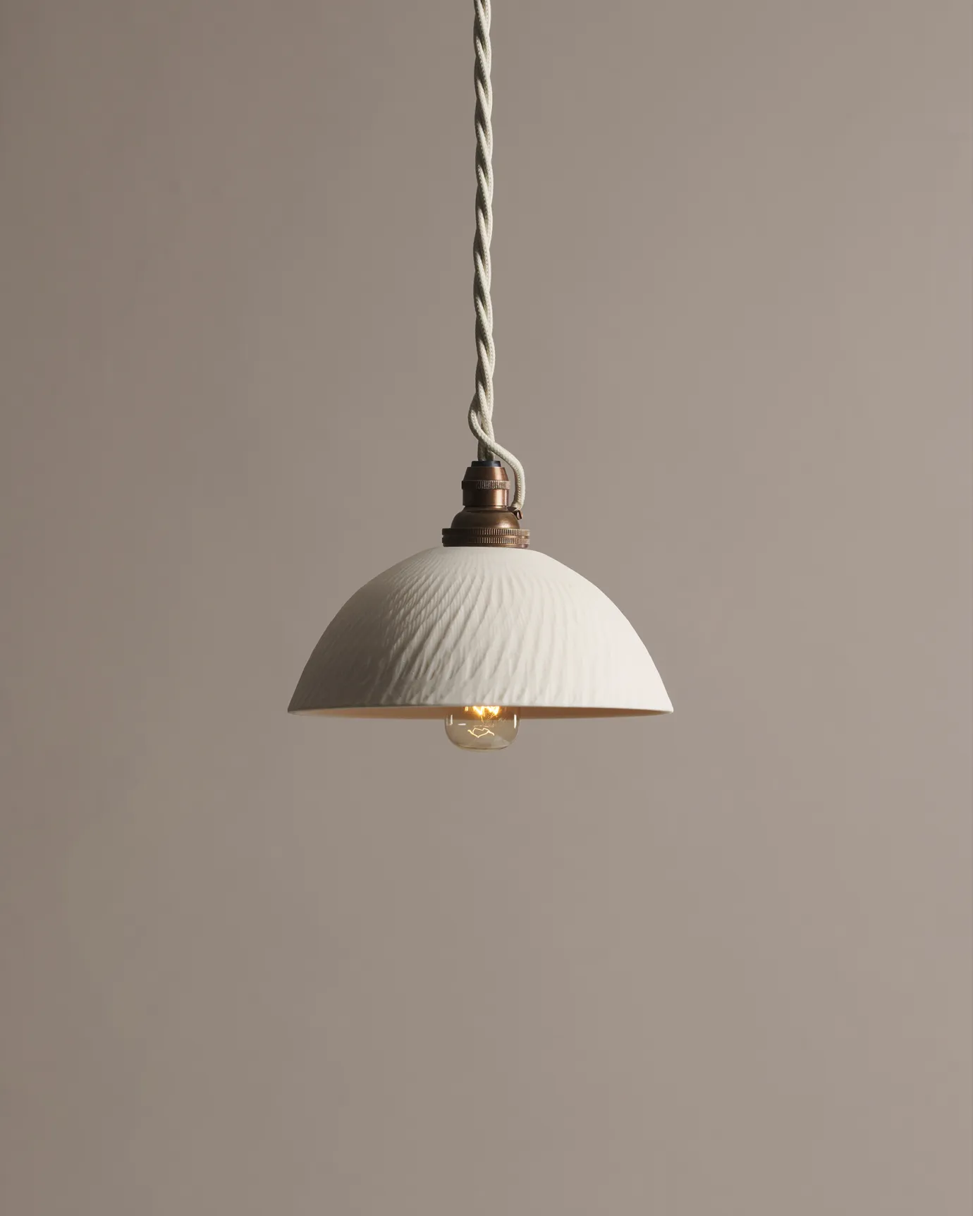 A chattered rice bowl porcelain pendant light with an unglazed finish, an aged brass B22 lampholder and a white twisted fabric flex cord.