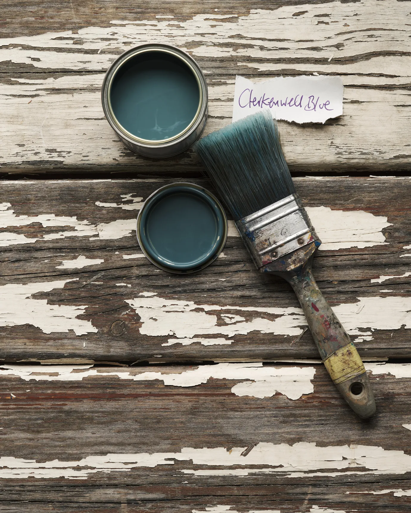 An overhead view of an open paint pot and a worn brush on a rustic tabletop. The green-tinged blue colour inside is labelled 'Clerkenwell Blue'.