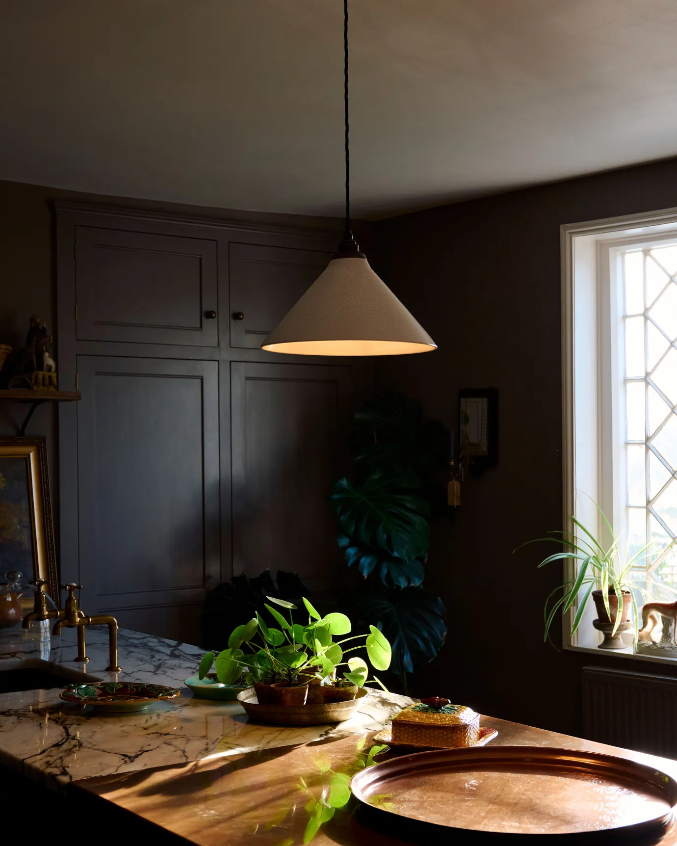 A white glazed ceramic pendant light with delicate crazing hanging in a dark room above an aged copper worktop.